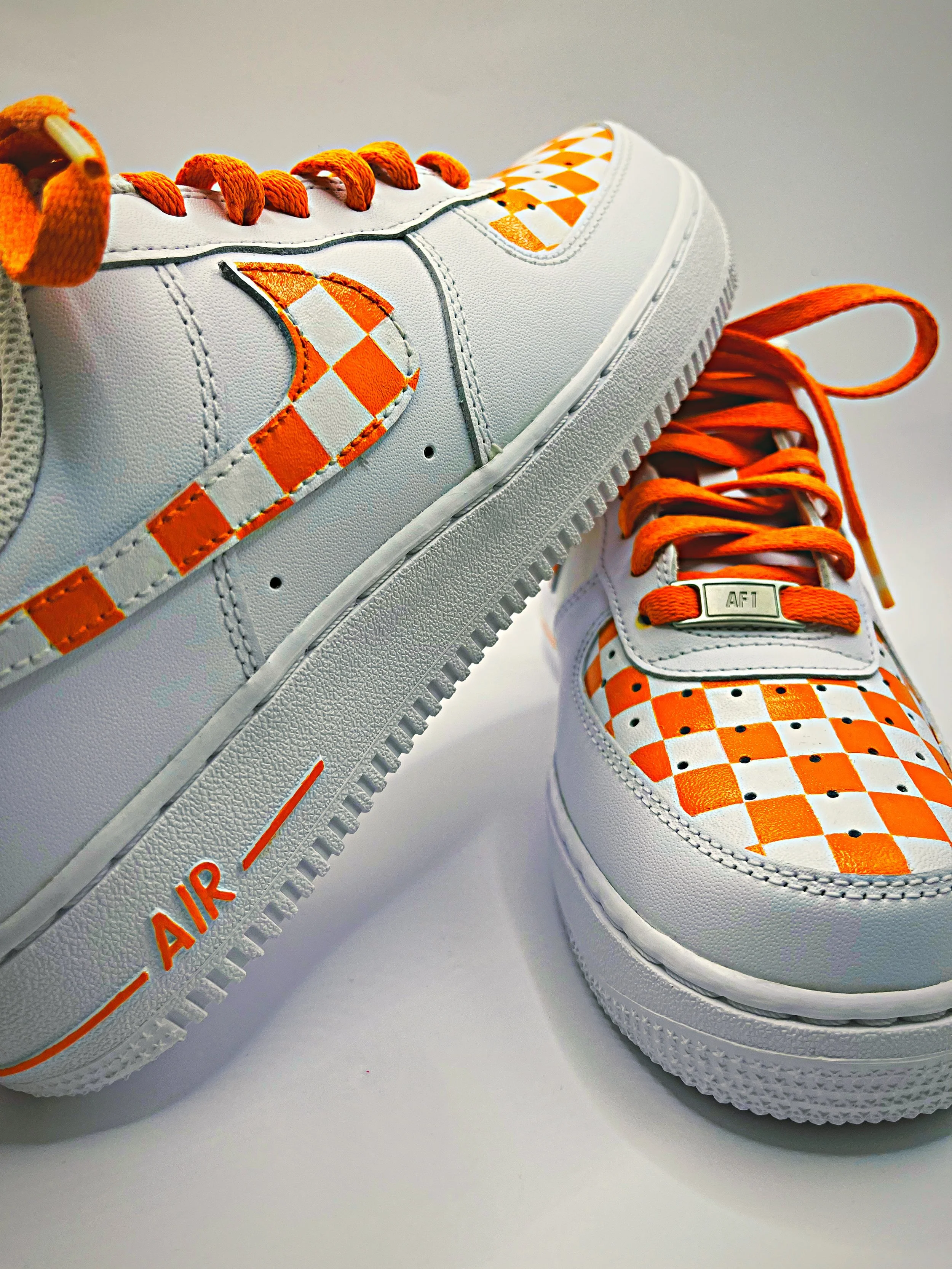 Close-up of a white sneaker with orange and white checkerboard pattern accents, orange laces, and an orange and white Nike swoosh logo.