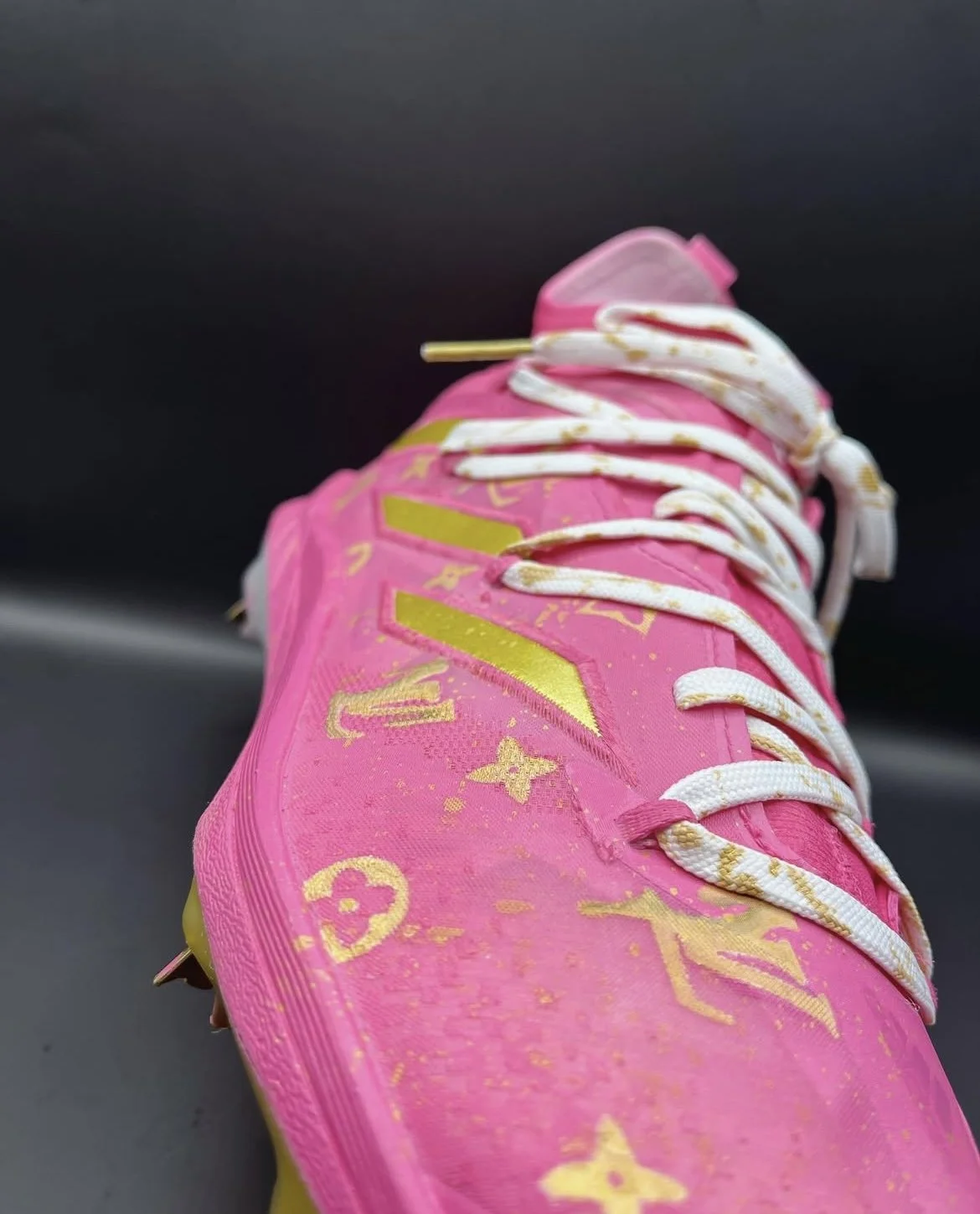 Close-up of a pink sneaker with gold markings and white laces, resting on a dark surface.