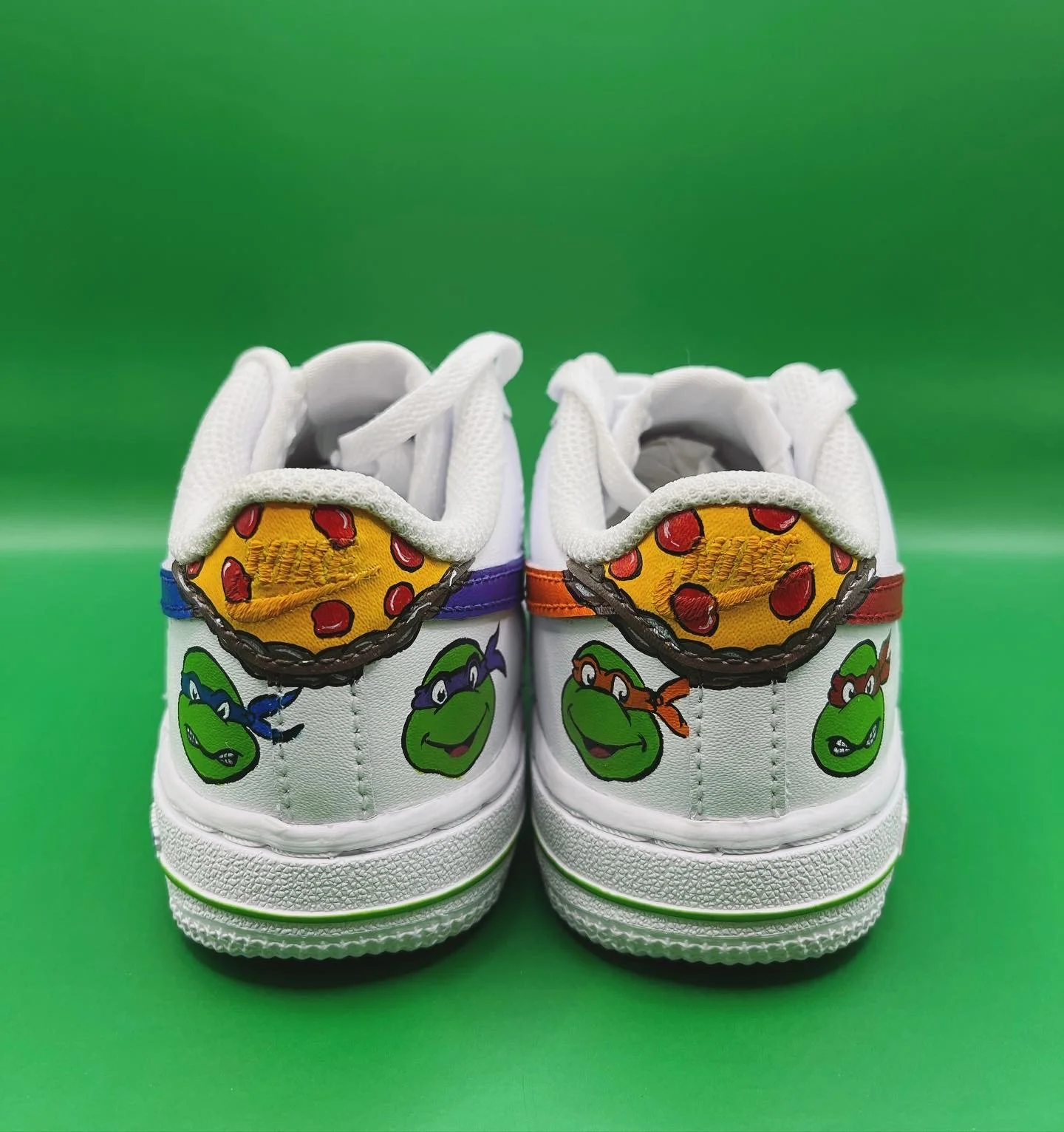 Pair of white sneakers with Teenage Mutant Ninja Turtles designs, featuring pizza and turtle faces, on a green background.
