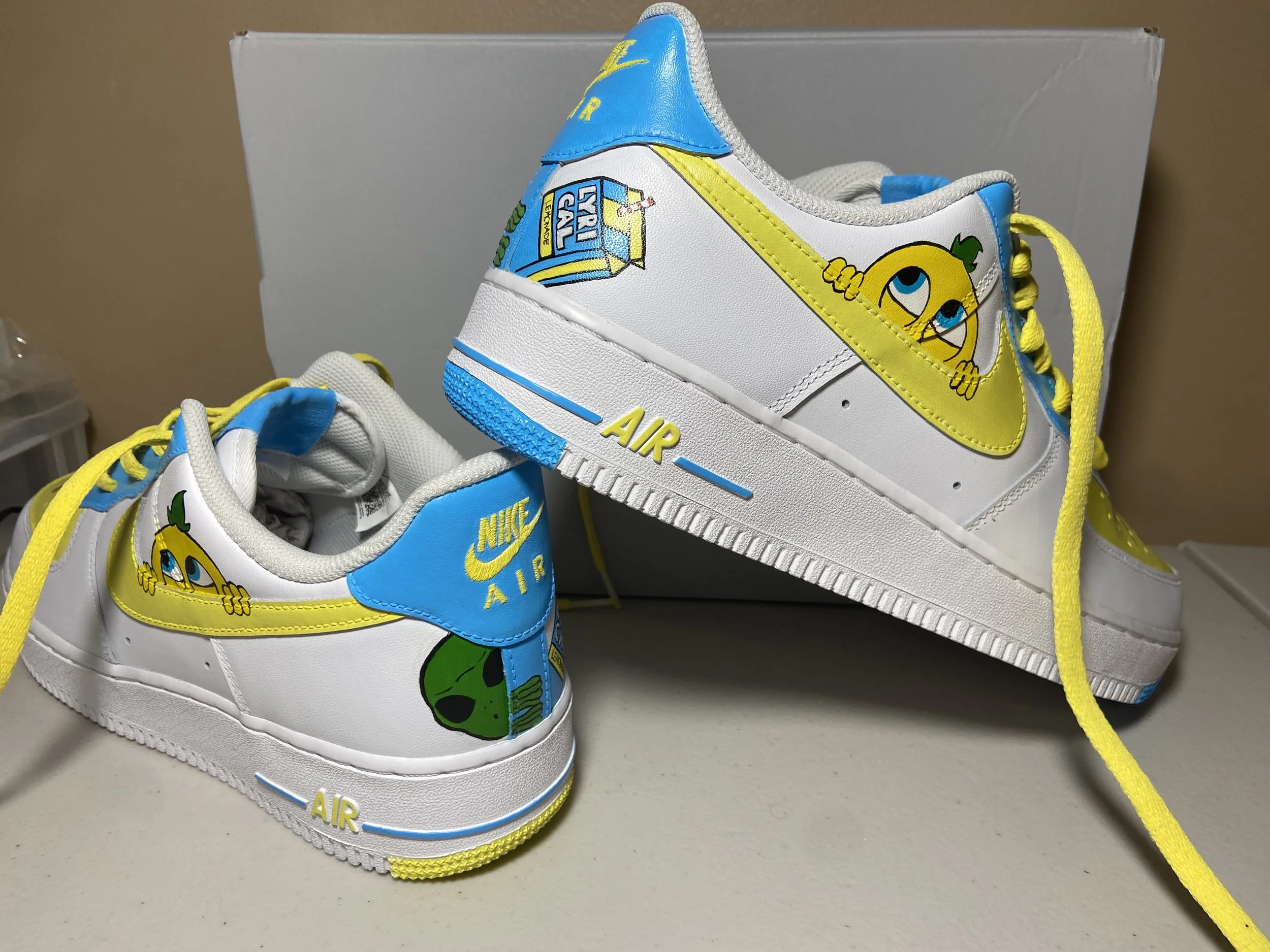 Pair of white Nike Air sneakers decorated with cartoon characters, including a yellow lemon with a green leaf and a blue-eyed girl, featuring colorful branding and accents in yellow, blue, and green.