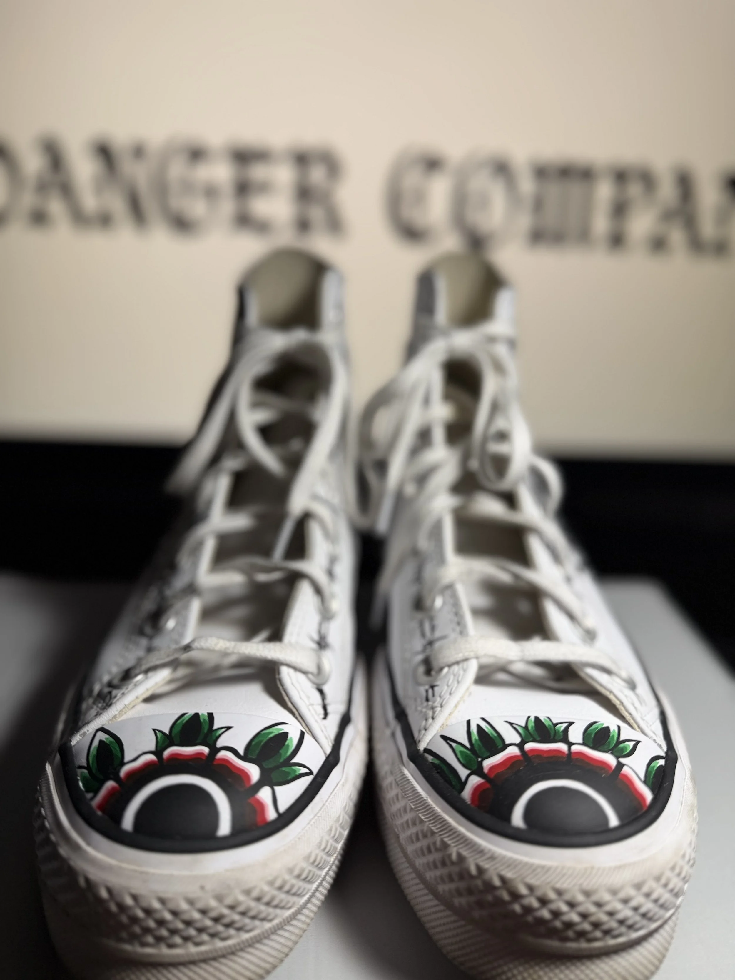 White sneakers with colorful floral designs on the toe caps, positioned in front of a beige background with blurred black text.
