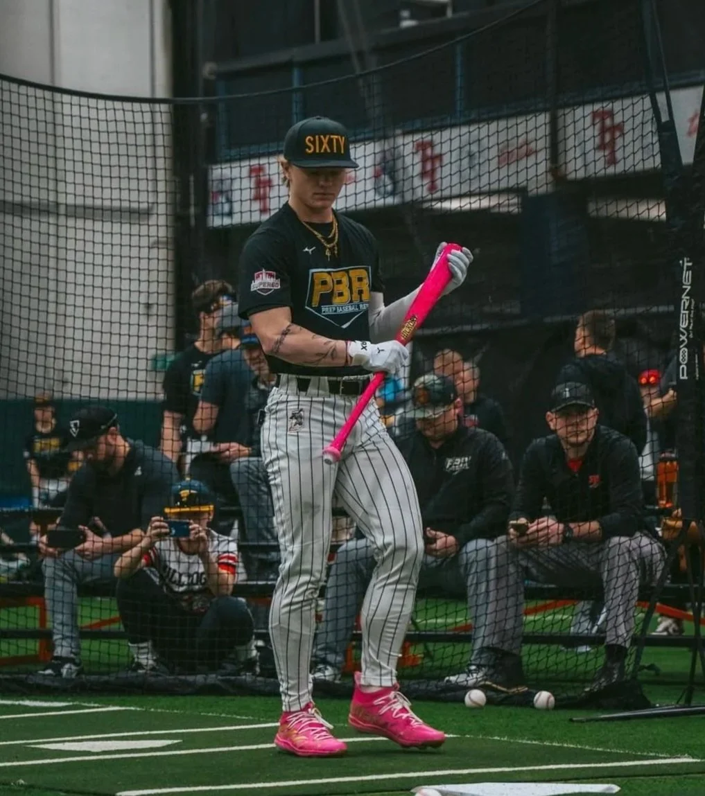 A young baseball player in pinstriped pants and a black t-shirt with PBR logo, wearing a black cap with 'SIXTY' on it, stands inside a batting cage holding a pink bat. Several people are seated and standing outside the cage, some using their phones.