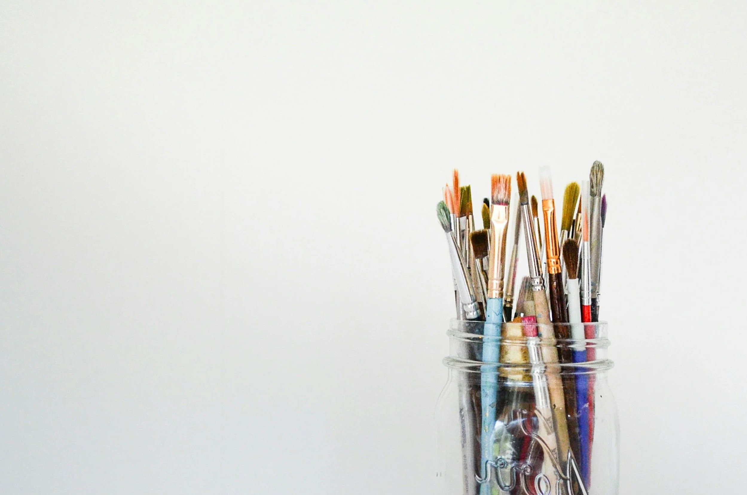 A glass jar filled with various paintbrushes of different sizes and styles, placed against a plain, light-colored background.
