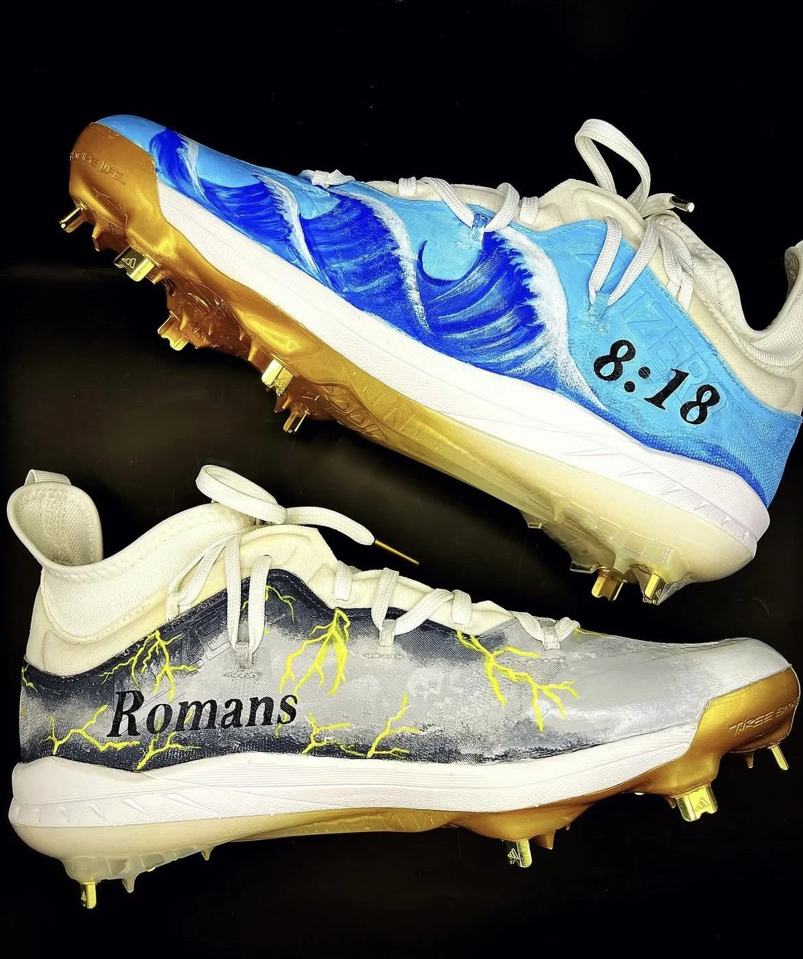 Two customized Nike baseball cleats with artistic designs. The top shoe has a wave pattern in blue and white with the date '8.18' written on the side, and the sole is gold. The bottom shoe features a lightning design in yellow and black with the word 'Romans' written on it and also has a gold sole.