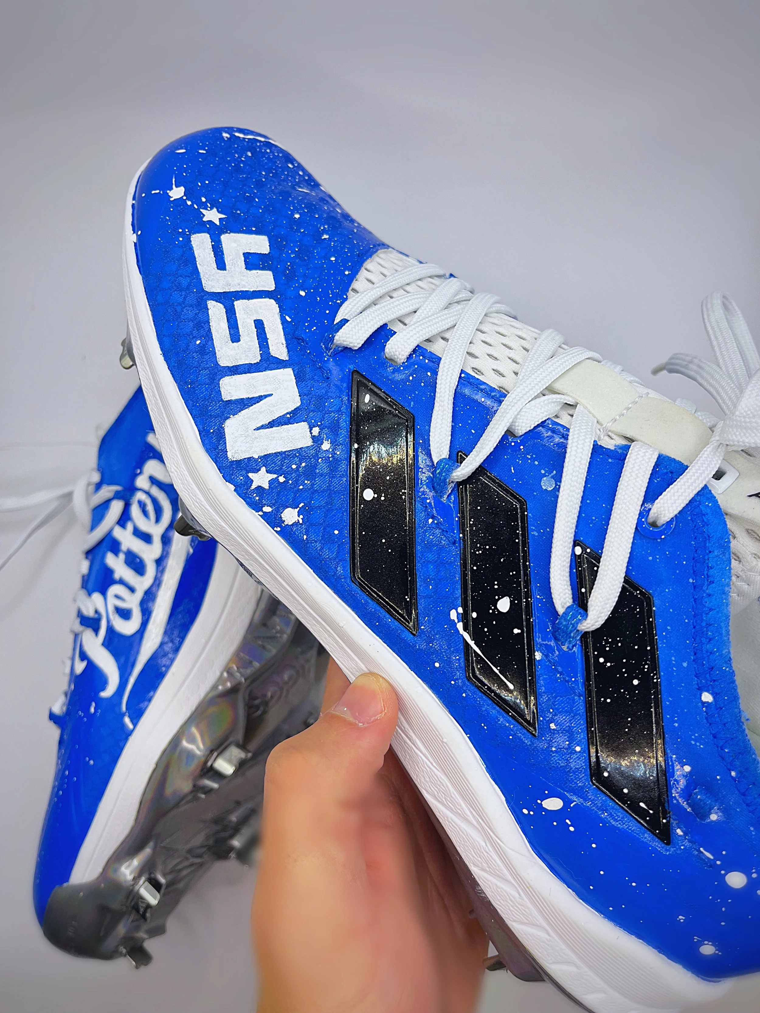 Close-up of a blue soccer cleat with white laces, white splatter paint design, and the word 'EVO' on the side, held by a person's hand.
