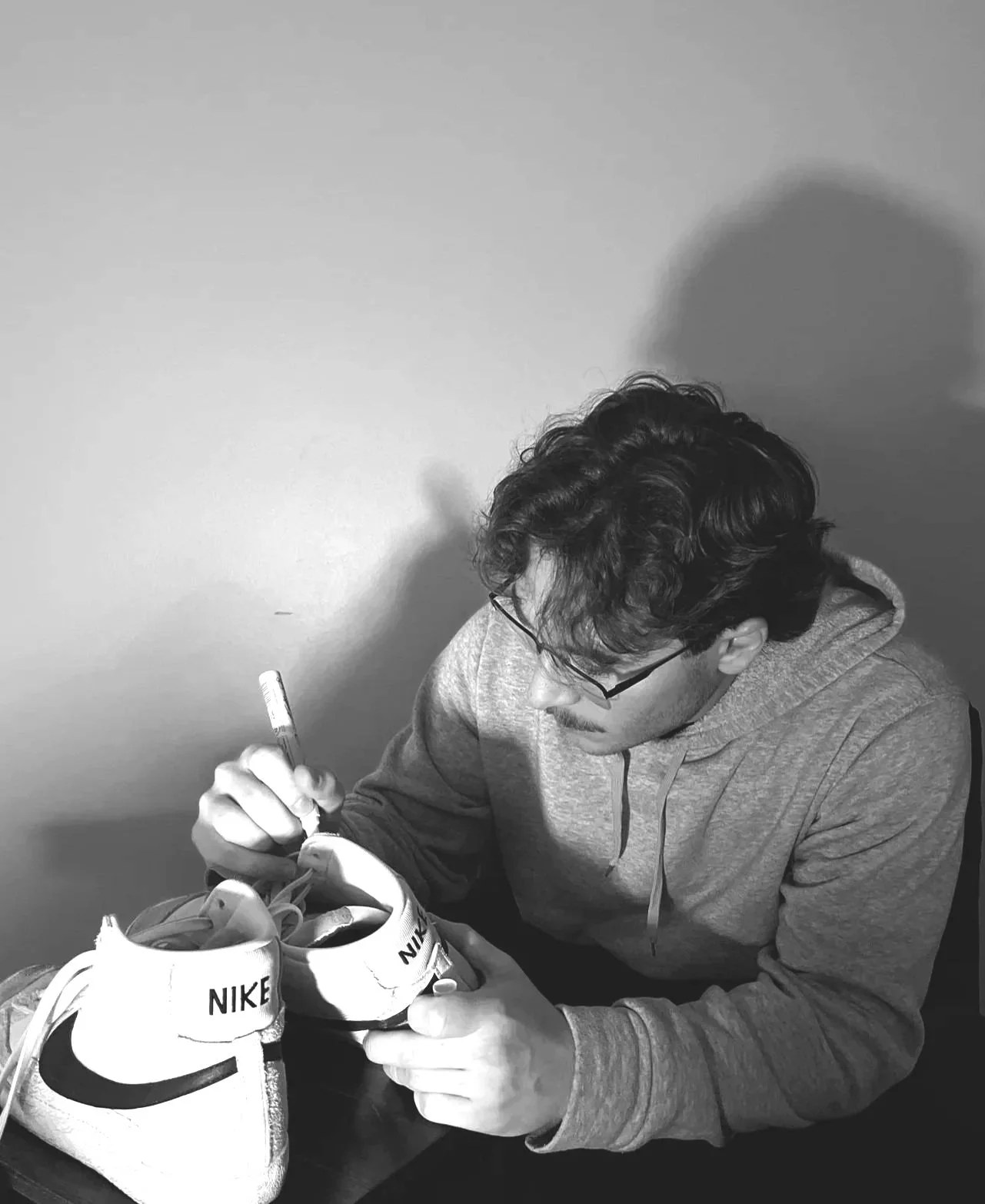 A man with glasses and dark curly hair sitting at a table, signing a pair of Nike sneakers.