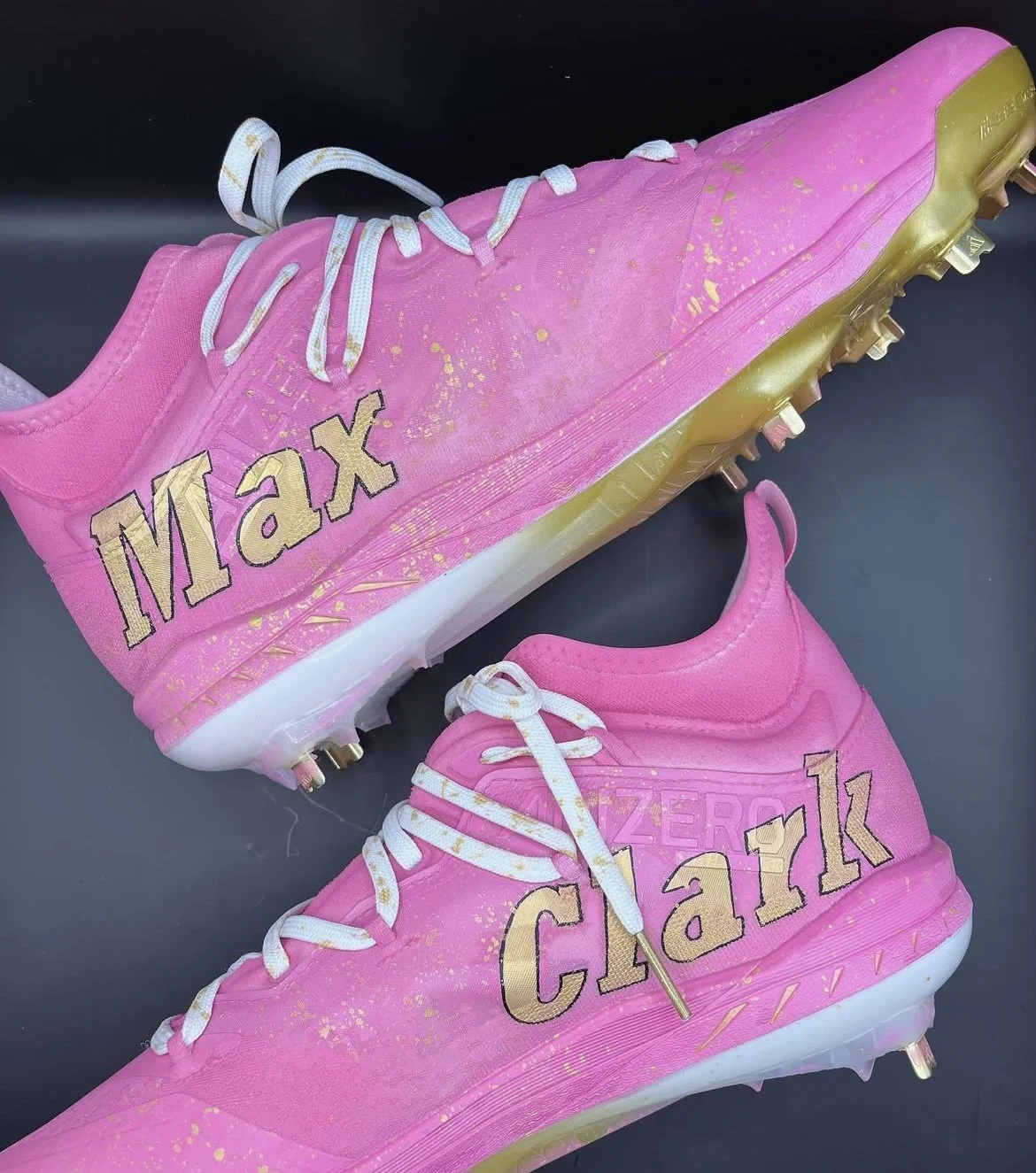Pair of pink baseball cleats with gold accents, labeled 'Max' and 'Clark', on a black background.