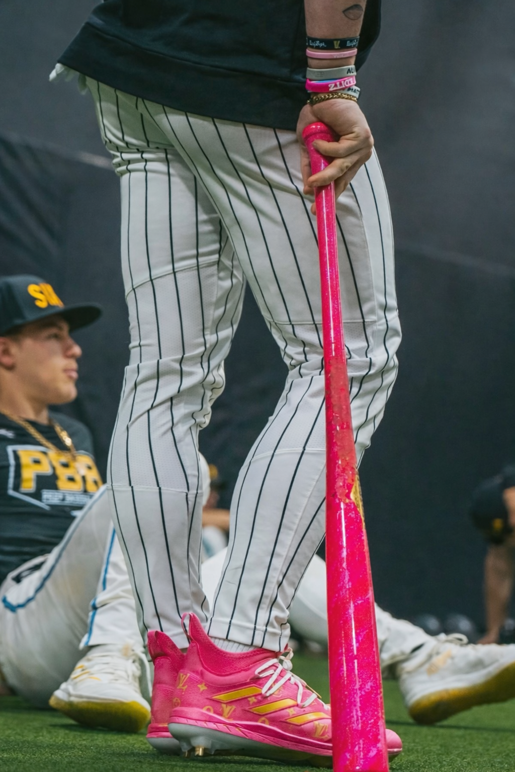 Close-up of a person wearing pink cleats and pinstripe baseball pants, holding a pink baseball bat, with a kneeling individual in the background.