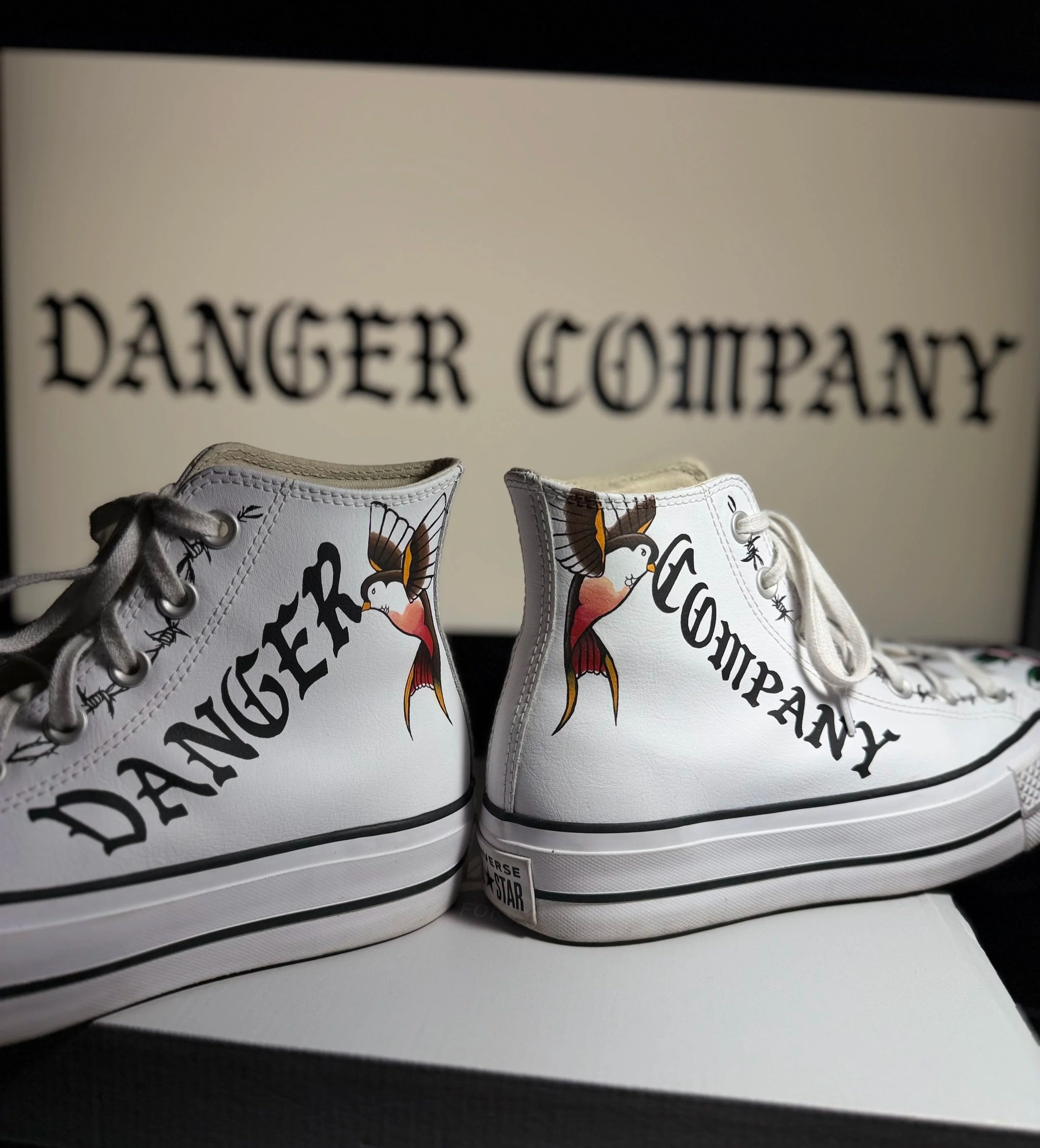 Close-up of white Converse sneakers with a butterfly and flamingo design, placed on a white surface, with a blurred background featuring text that reads 'DANGER COMPANY'.