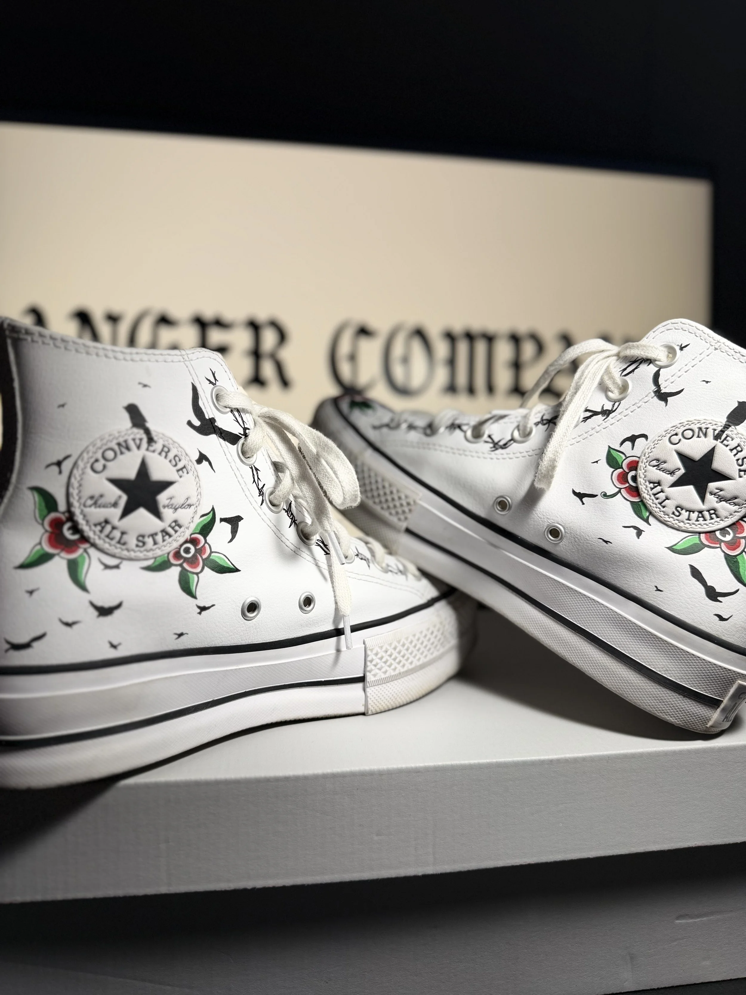 Pair of white Converse Chuck Taylor All Star sneakers with a floral and bird design, placed on a box with a blurred beige and black background.