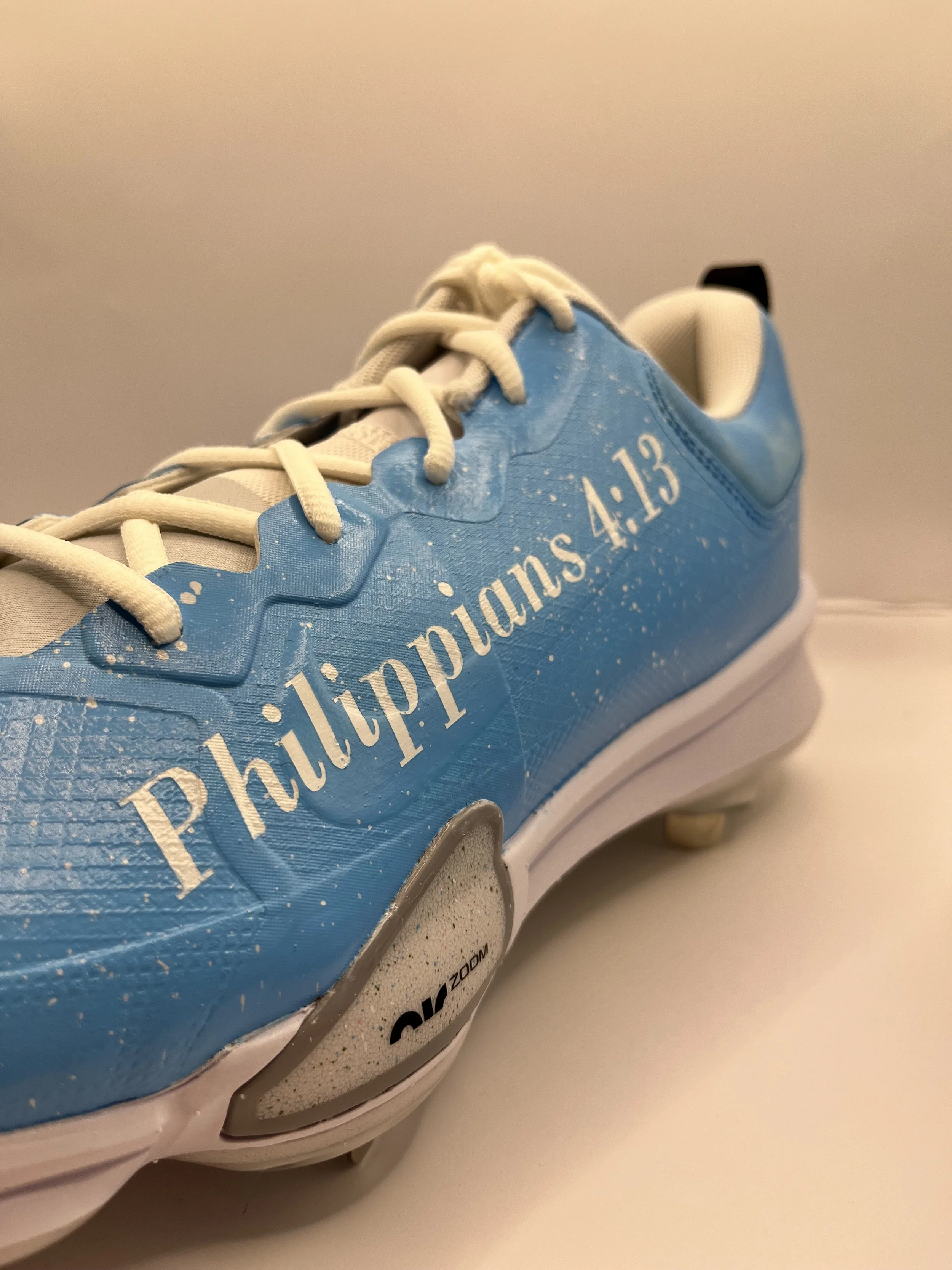 Close-up of a blue athletic shoe with white laces, featuring white text that reads "Philippians 4:13," resting on a white surface.