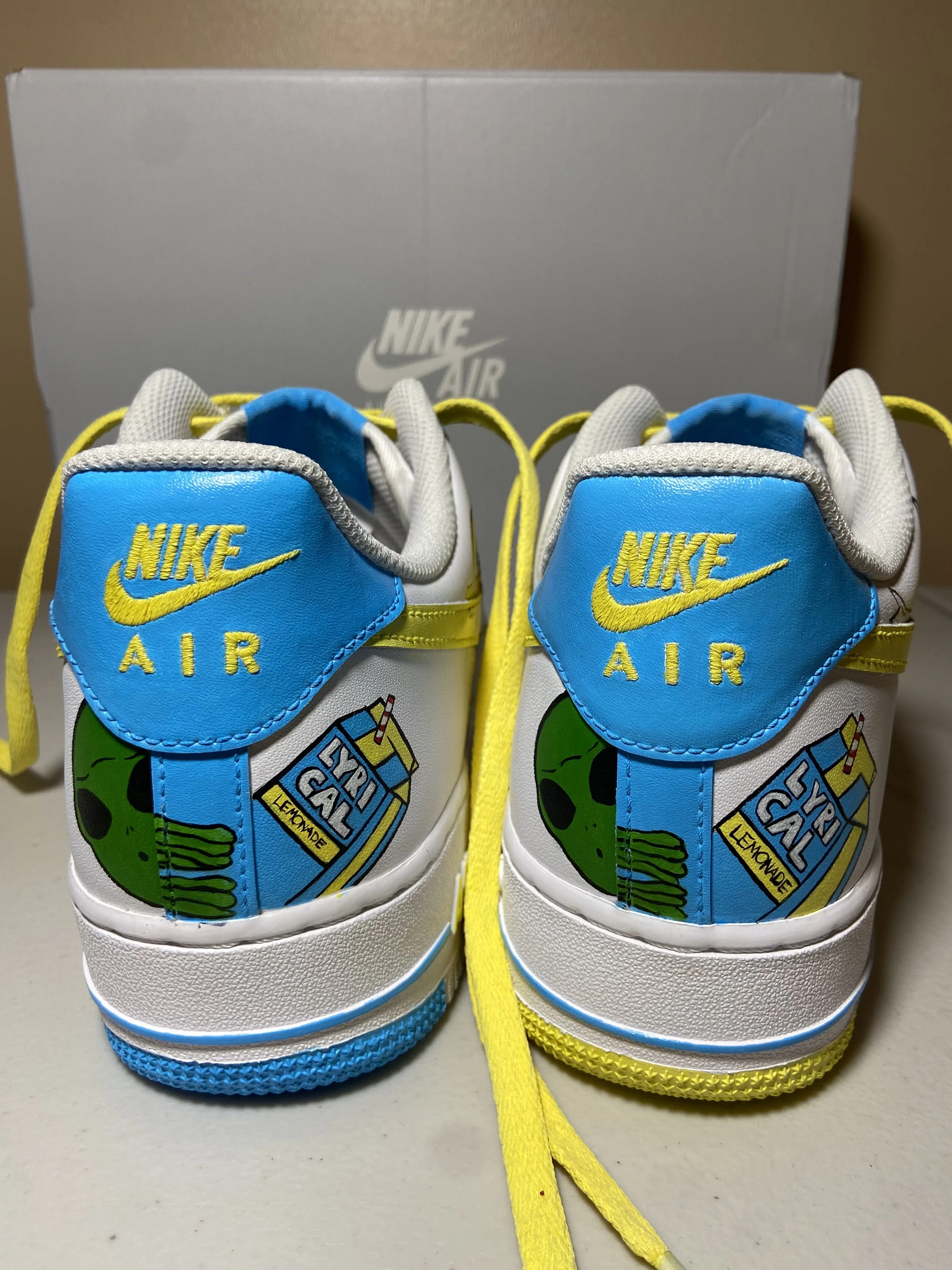 Close-up of a pair of white Nike Air sneakers with cartoonish designs on the back and yellow shoelaces, with a Nike shoebox in the background.
