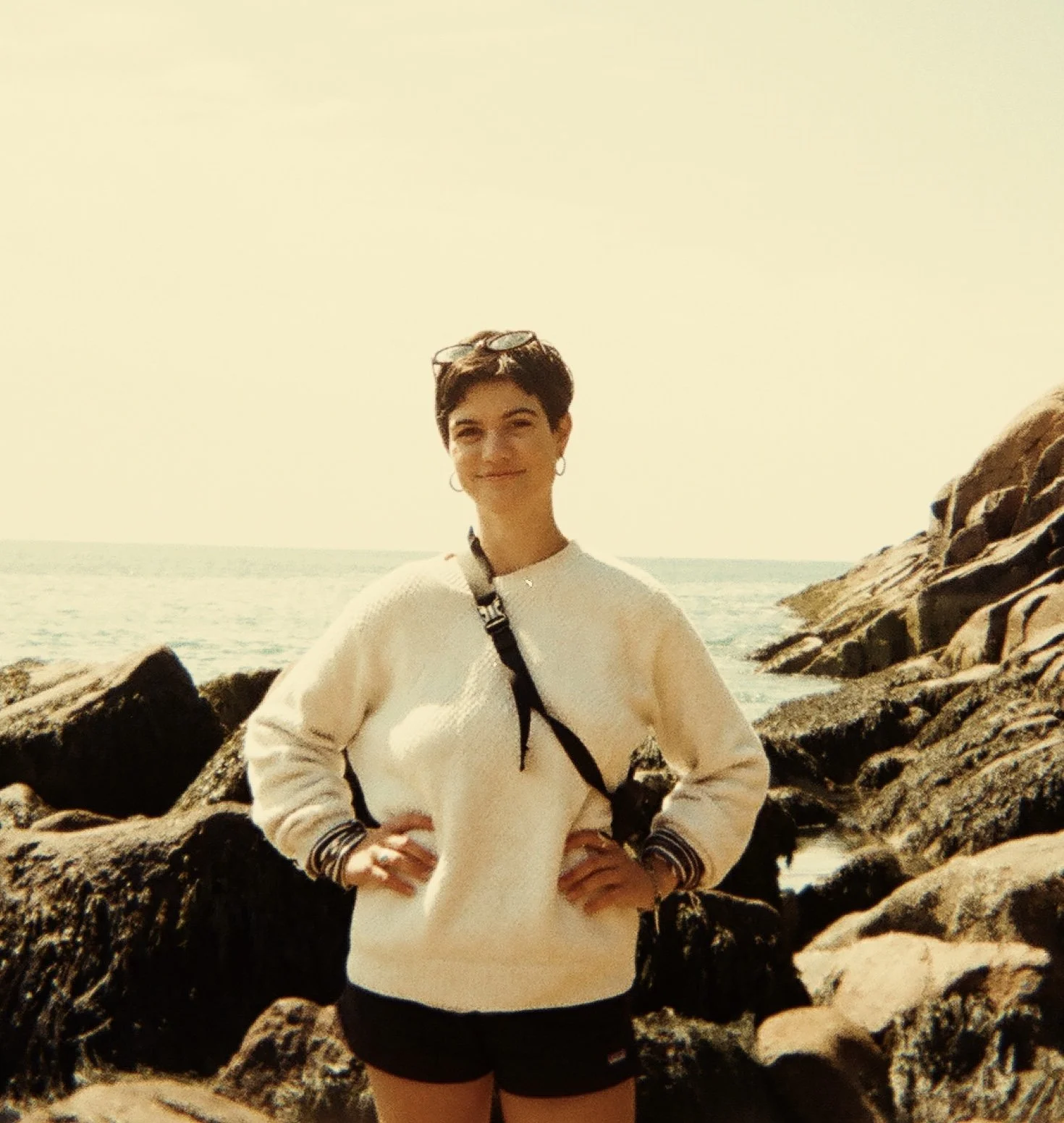 Woman standing on rocks at the beach with ocean in the background, wearing a cream sweater, black shorts, and sunglasses on her head, smiling with hands on hips.