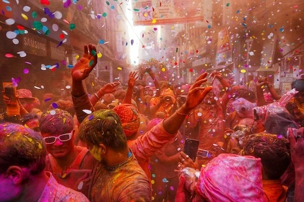 A crowd of people celebrating during a Holi festival, covered in colorful powders with confetti in the air.