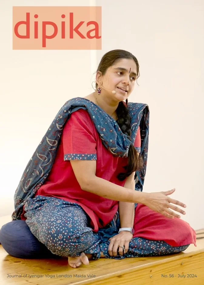 A woman sitting cross-legged on a wooden floor, wearing a pink top with blue patterned pants and a matching blue shawl, speaking into a microphone headset. There is a title 'dipika' in red text at the top left corner, and smaller text at the bottom mentioning the Journal of Iyengar Yoga London Maida Vale, issue number 56 from July 2024.