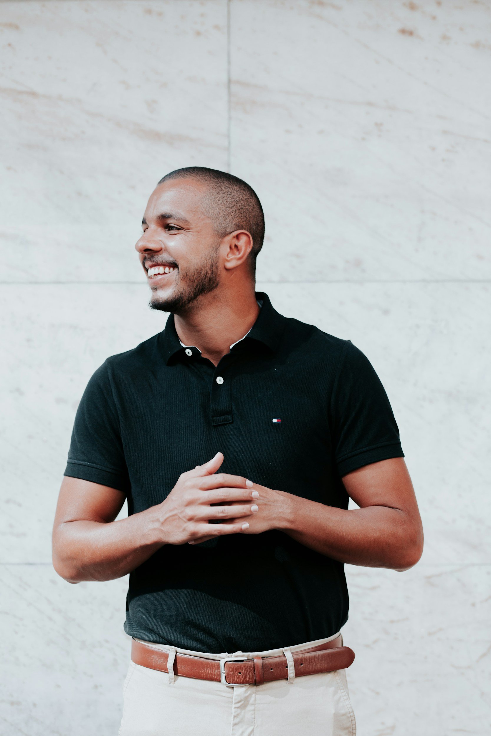 A smiling man with a beard, wearing a black polo shirt, white pants, and a brown belt, standing against a light-colored wall.