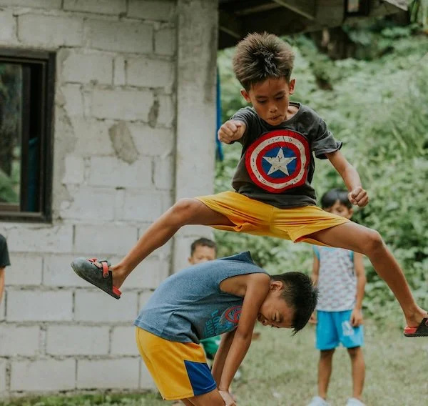 A boy jumps high over another boy who is bending down, with a background of a house and greenery.