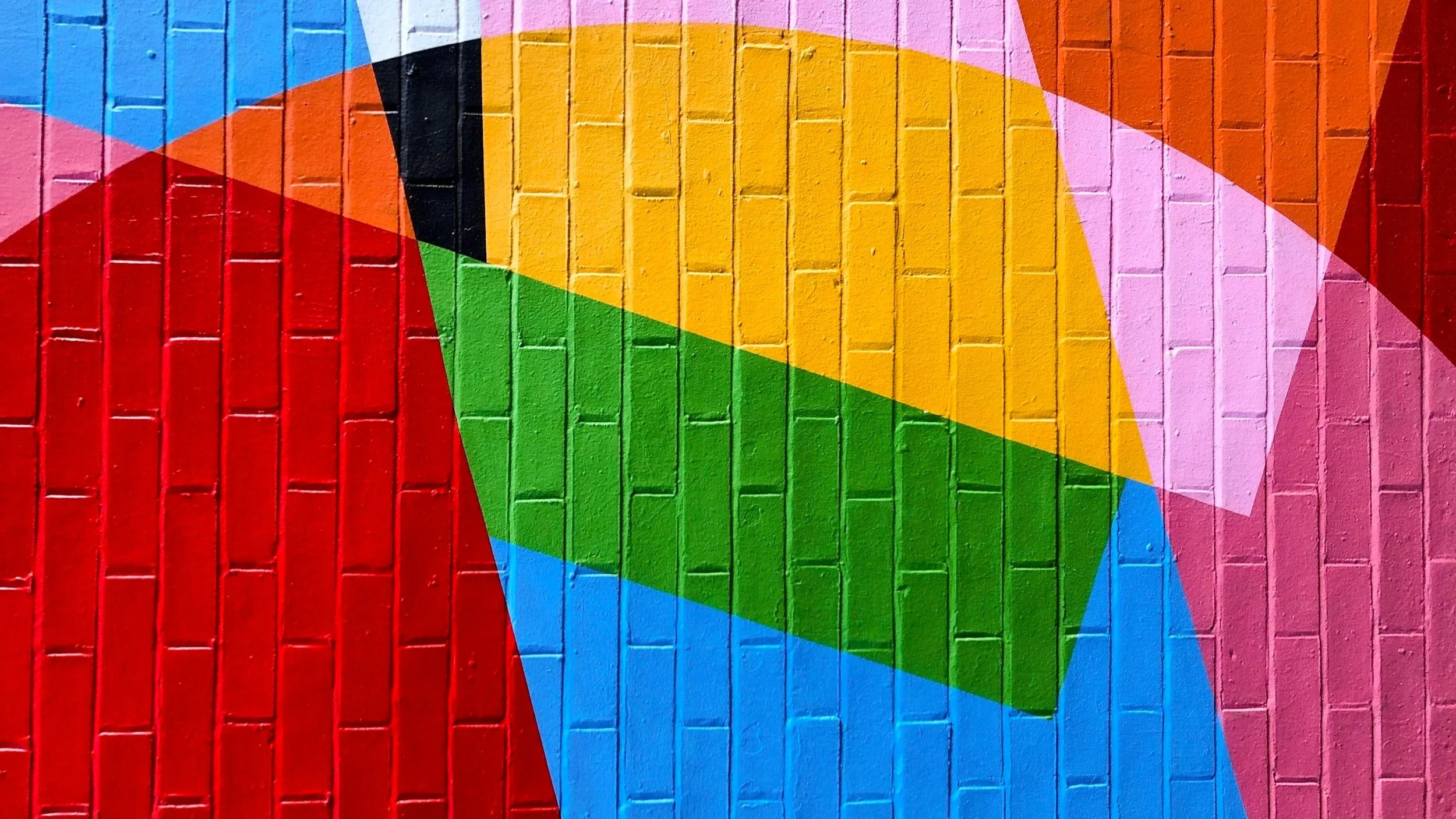 Colorful abstract mural painted on a brick wall with overlapping geometric shapes in red, blue, green, yellow, pink, black, and white.