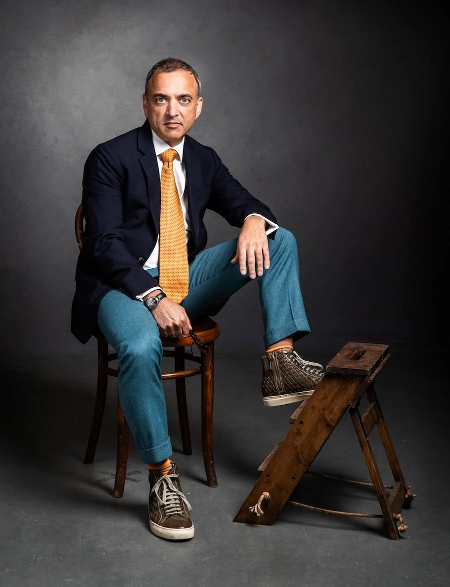 A man seated on a wooden chair with one foot raised on a small wooden step stool, wearing a navy blazer, orange tie, white shirt, blue pants, and sneakers, against a dark, plain background.