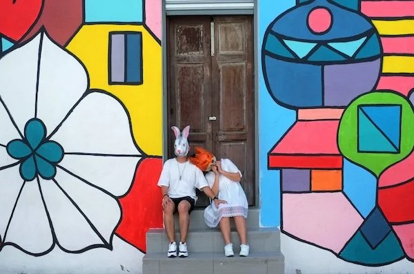 Two children sitting on a step outside a building with colorful abstract mural art, one wearing a bunny mask and the other with an orange object on their shoulder.