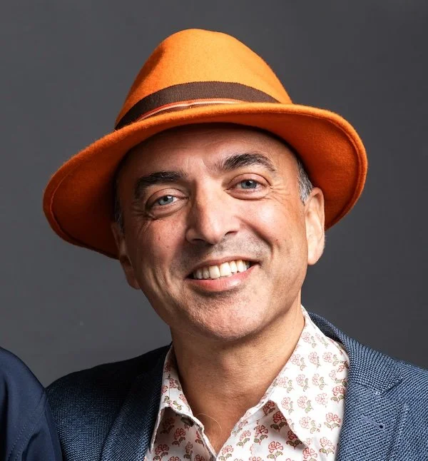 A man wearing an orange fedora hat, a white patterned shirt, and a blazer, smiling at the camera against a dark background.