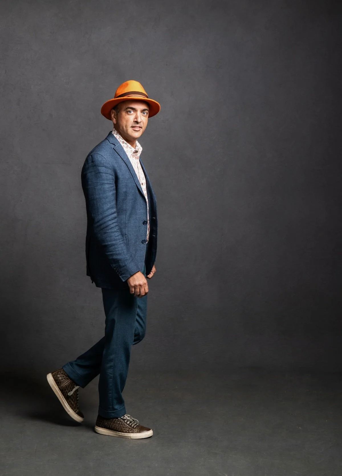 A man in a gray suit, patterned shirt, and brown sneakers standing against a dark gray background, wearing a yellow fedora hat.