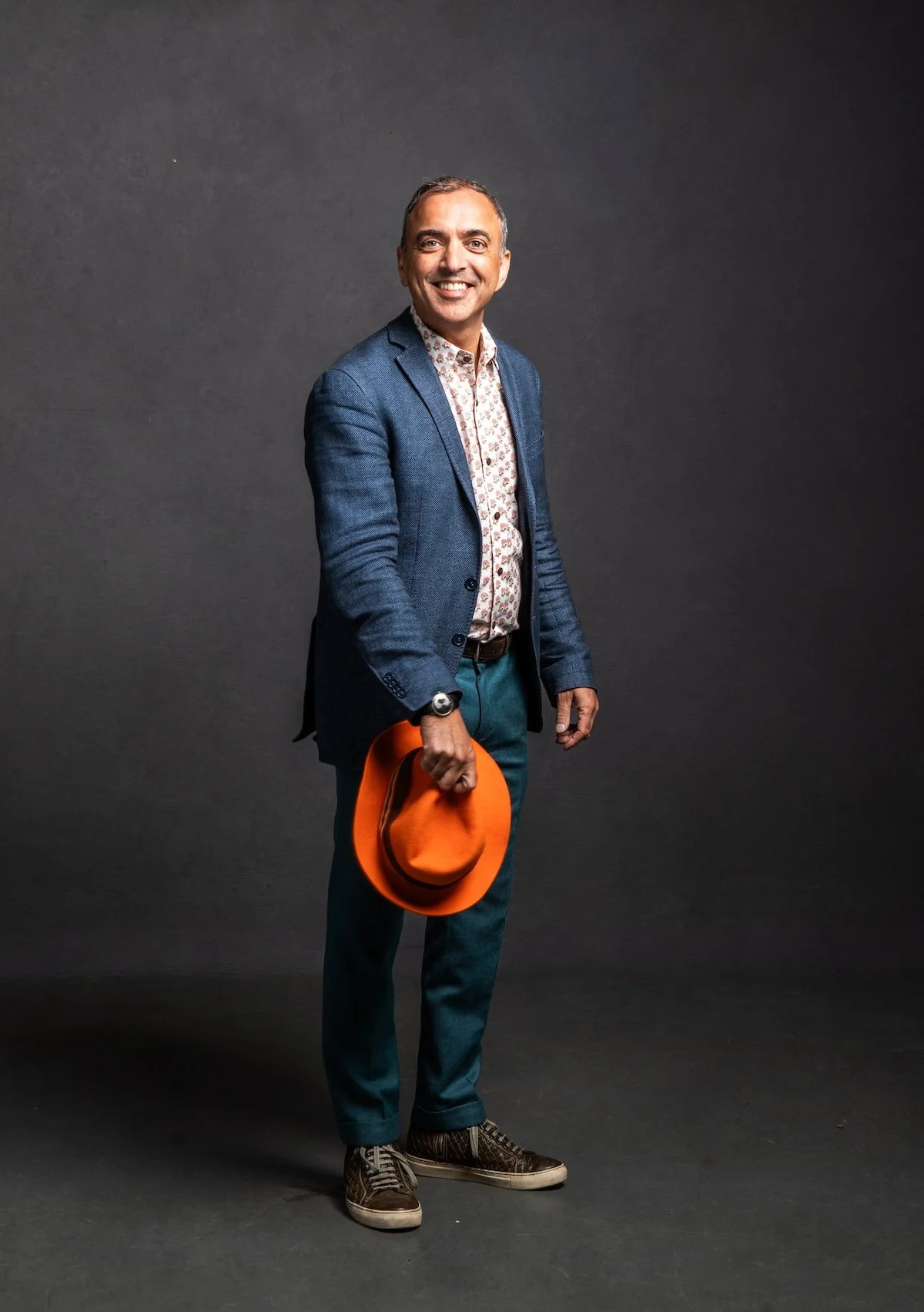 A smiling man in a blue blazer, patterned shirt, and green trousers holding an orange hat standing against a dark gray background.