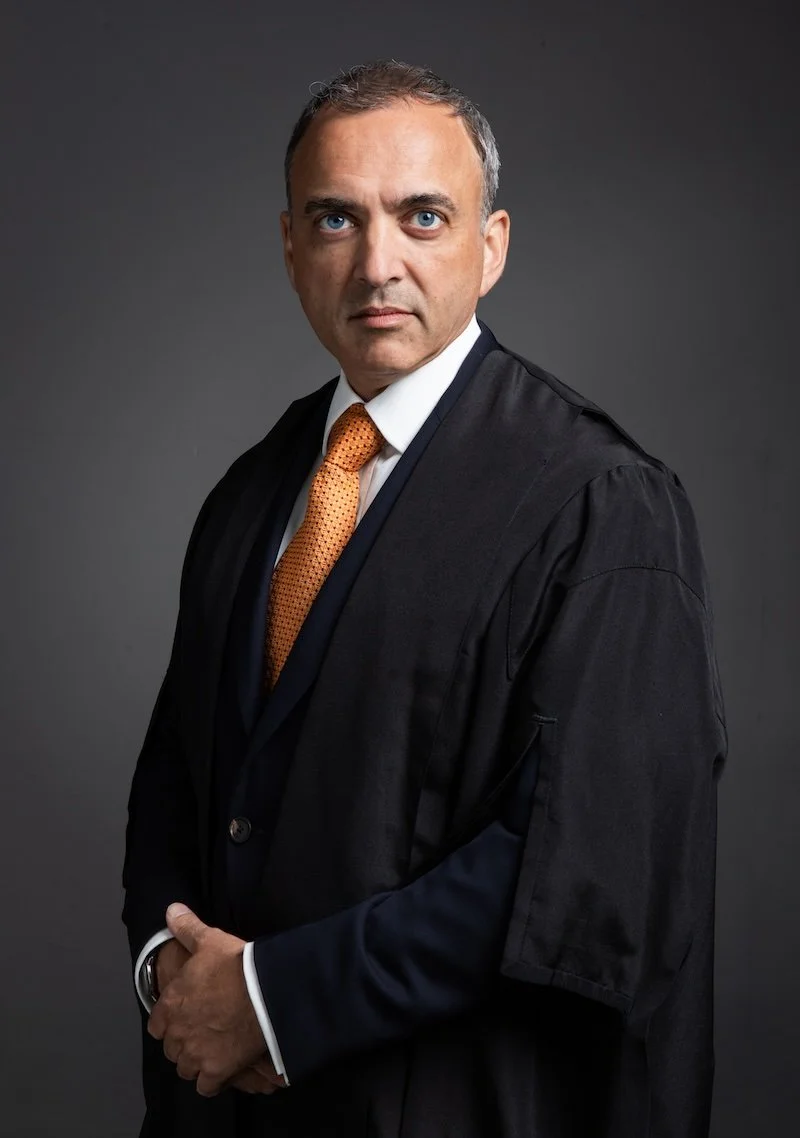 Professional man in a black robe and orange tie posing against a dark gray background.