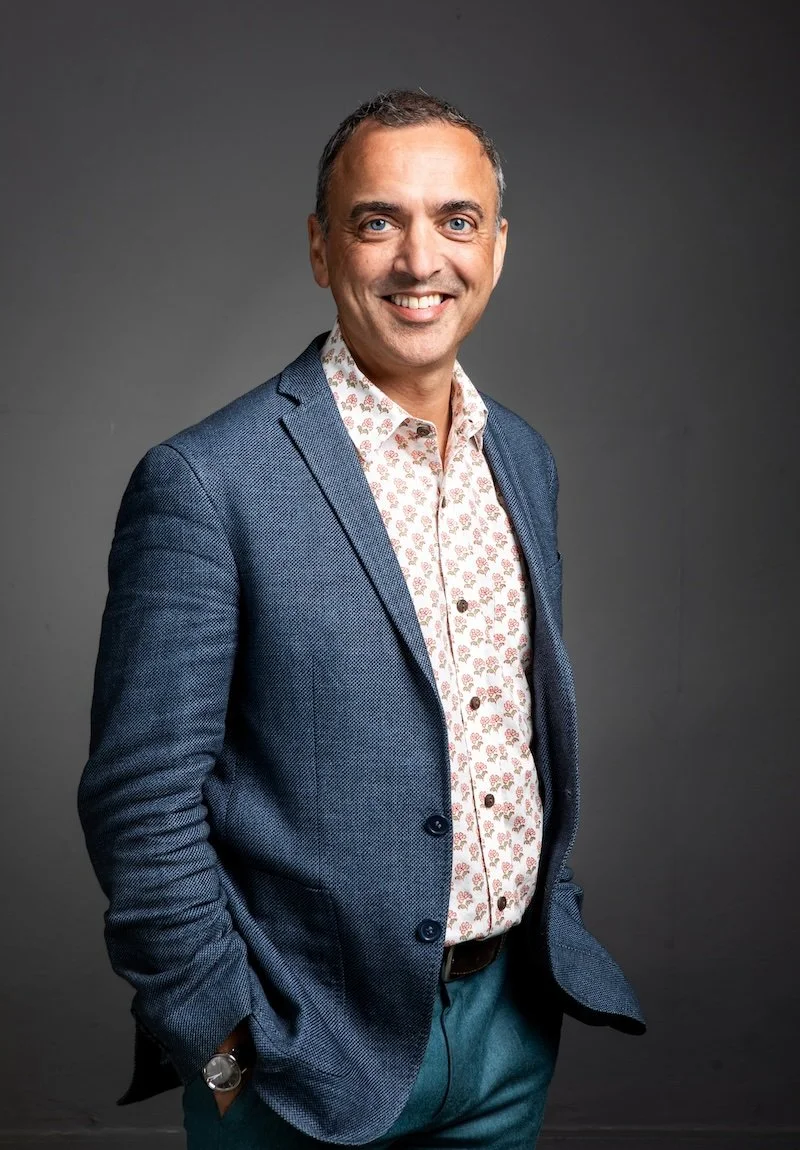 A smiling man with blue eyes and short dark hair, wearing a blue blazer over a white shirt with a pink floral pattern, standing against a dark gray background.