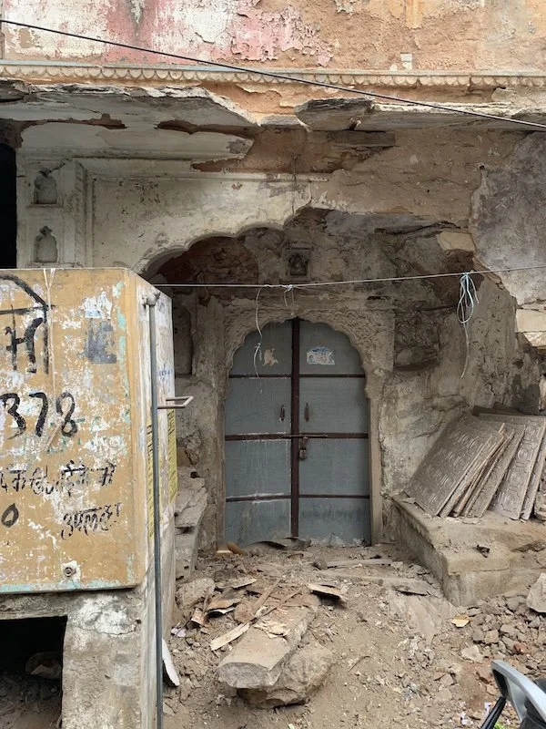 Old metal door set into an excavated wall with crumbling plaster and debris on the ground, surrounded by construction materials and exposed brick.