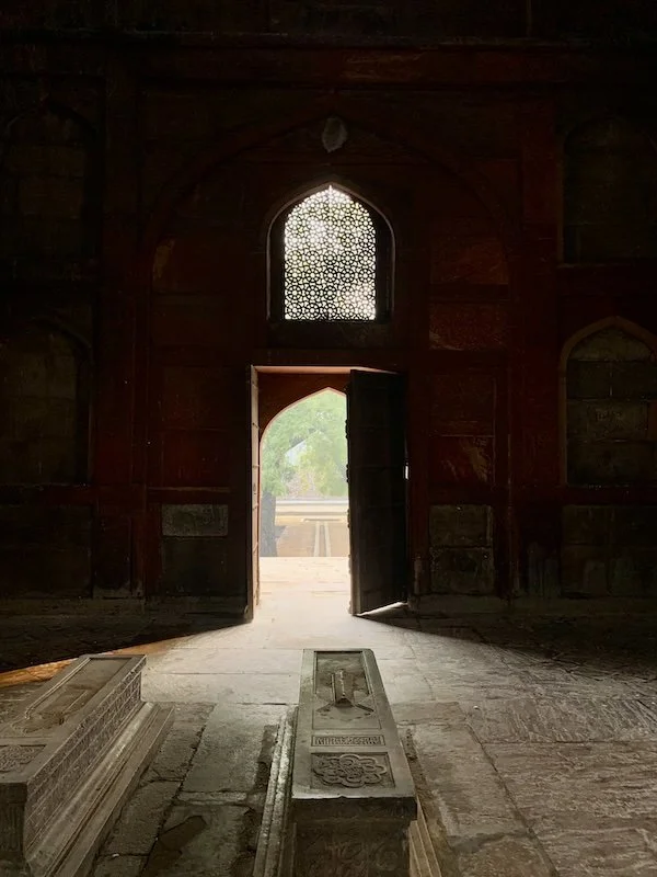 The interior of an old building with a high arched doorway open to a balcony or courtyard outside. Sunlight streams through, revealing intricate patterns in the window above the door. On the floor inside, there are carved stone or marble slabs, possi