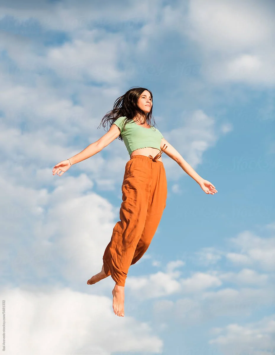 A woman appears to be levitating in mid-air against a blue sky with clouds, with her arms outstretched and her eyes closed.