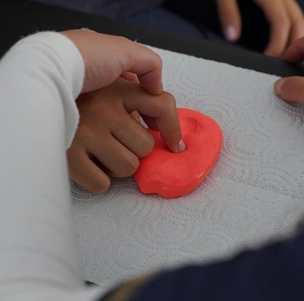 A child's finger pressing into a piece of pink modeling clay on a white paper towel.