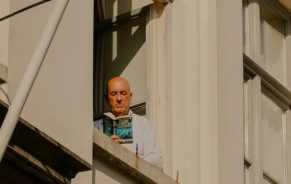 A man reading a book on a balcony with sunlight and shadows.