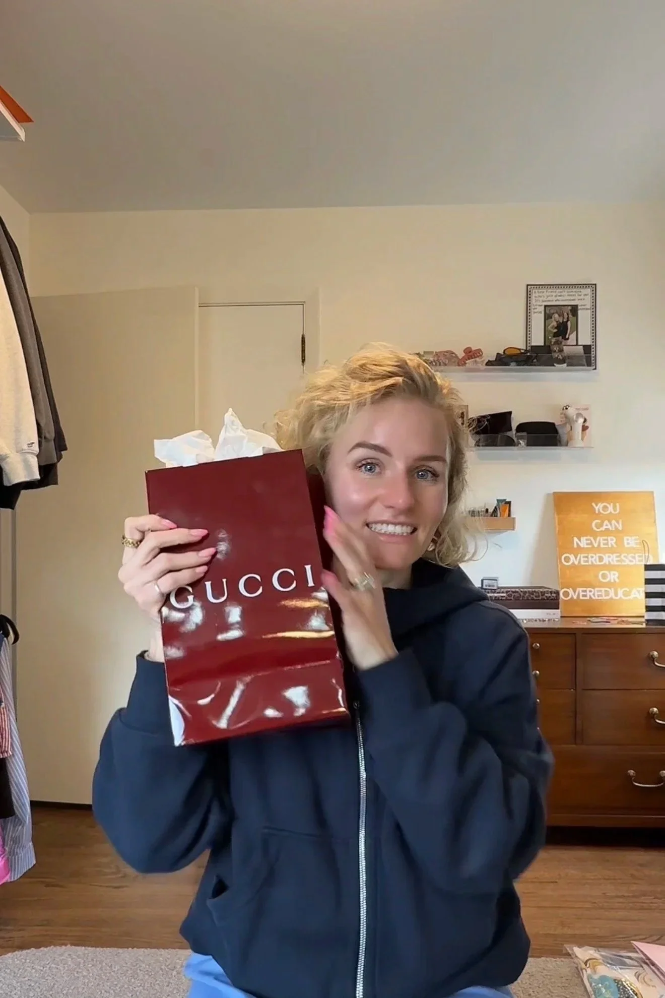 A woman with blonde, curly hair smiling and holding a red Gucci shopping bag. She is wearing a black hoodie. Behind her is a wooden dresser and shelves with various items. There is a framed picture and a sign that reads, 'You can never be overdressed or overeducated.'