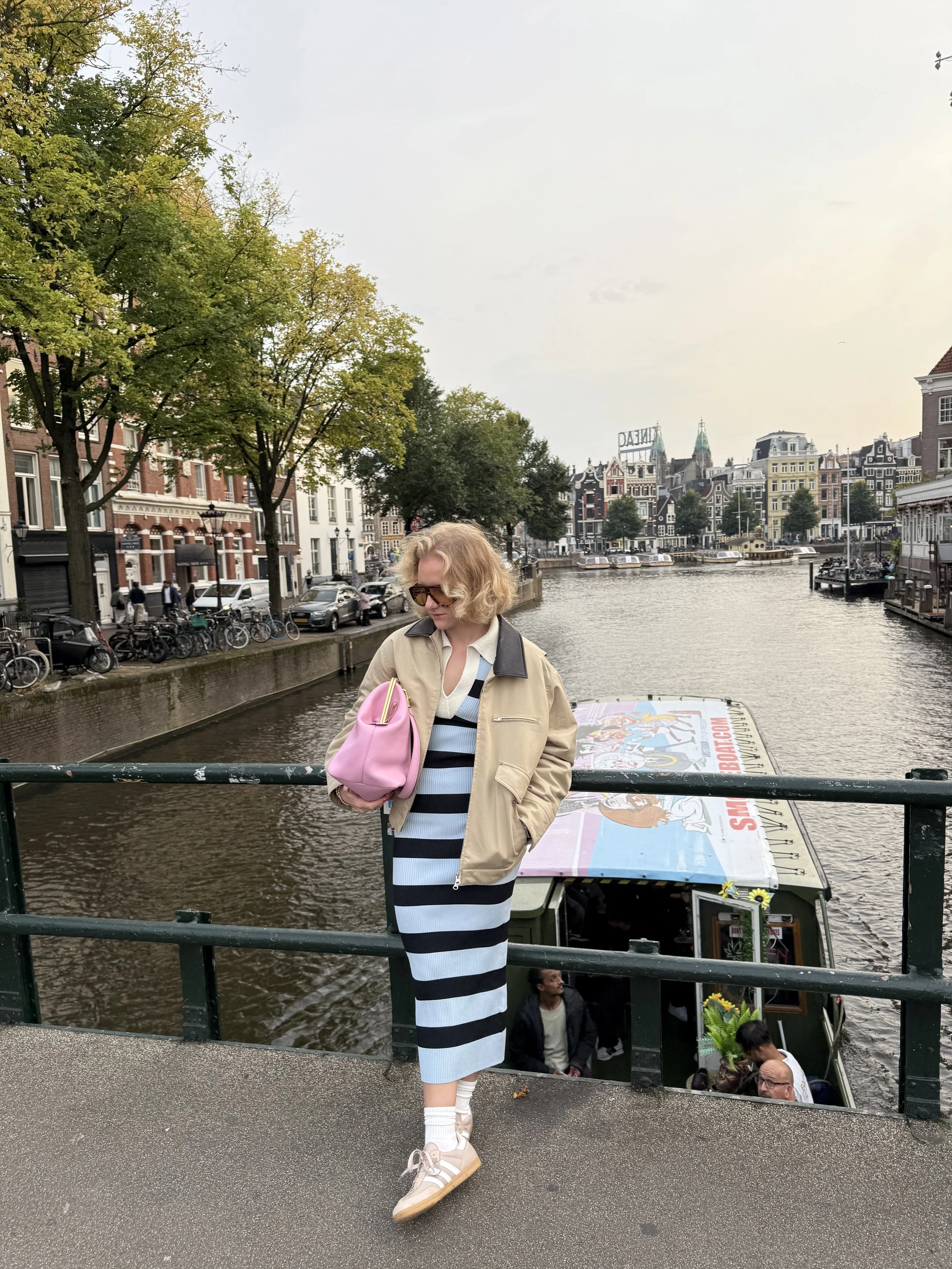 A woman with curly blonde hair, wearing sunglasses and a beige jacket, stands on a bridge over a canal in a European city. She is holding a pink bag and wearing a striped blue and black dress with white sneakers. In the background, there are buildings, trees, parked cars, boats on the canal, and a sign that reads 'FINLAND' on top of a building.