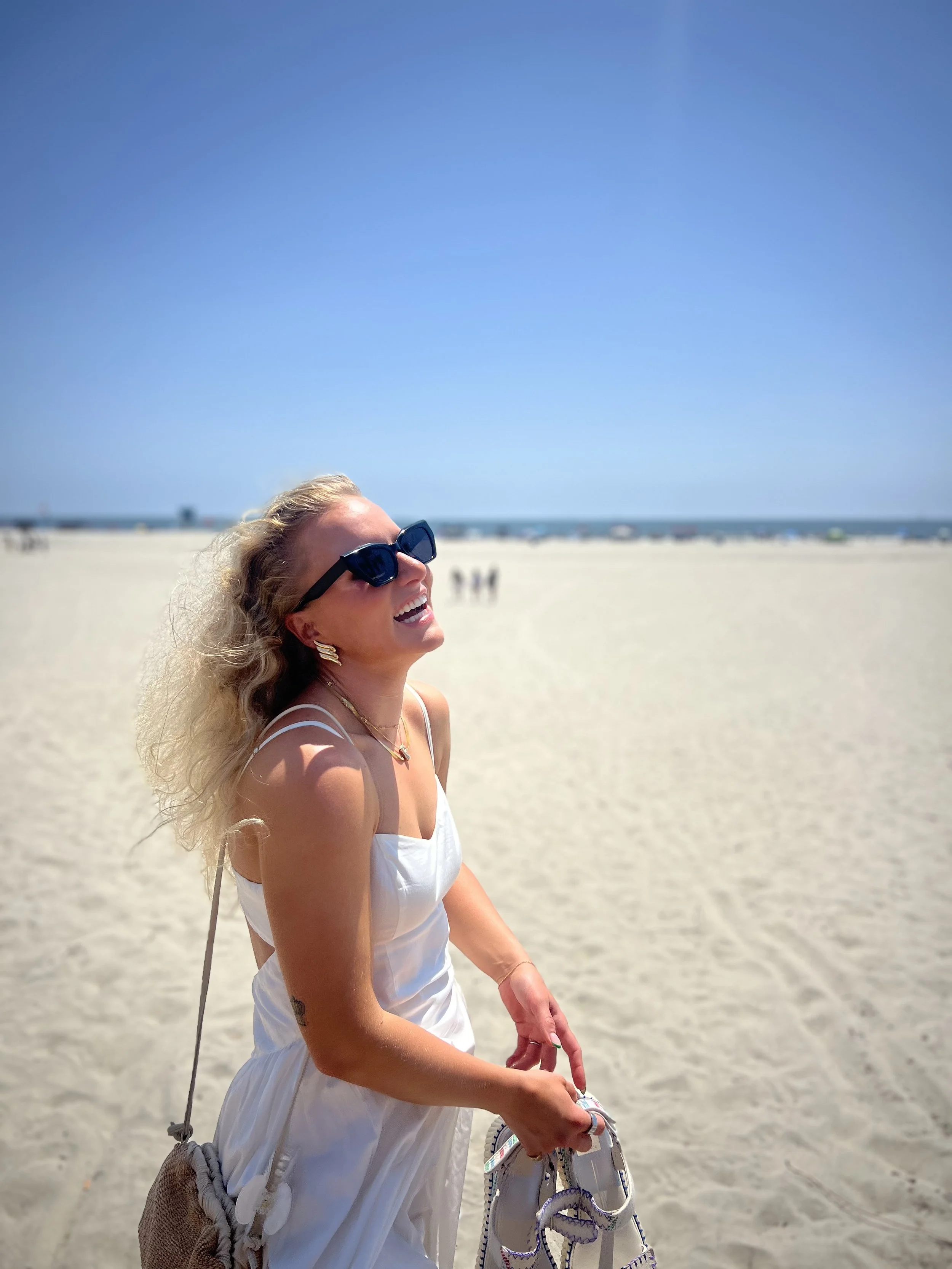 A woman with curly blonde hair wearing sunglasses, a white dress, and earrings standing on a sandy beach with a clear blue sky above and a few people in the background.