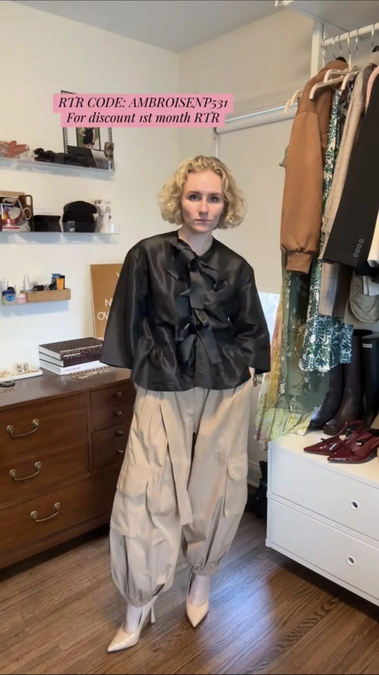 A woman with curly blonde hair wearing a shiny black top with a bow, loose beige cargo pants, and white high-heeled shoes standing in front of a dresser and a clothing rack in a room.
