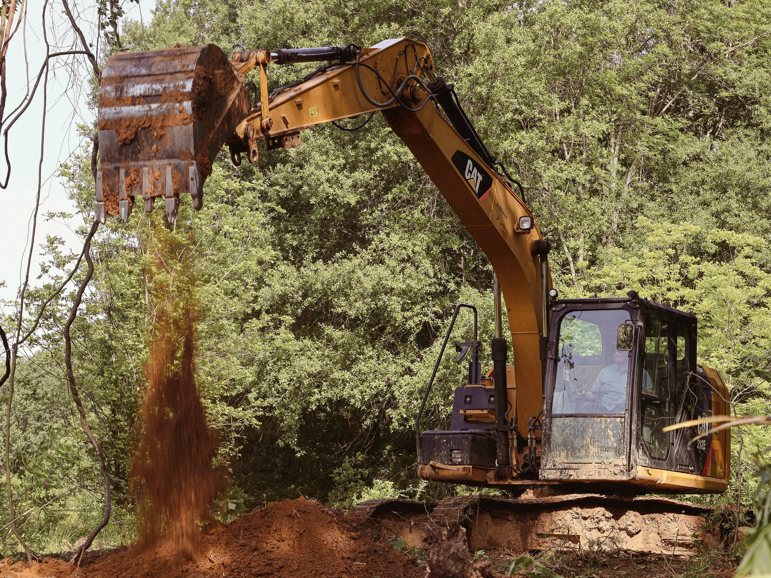 A large construction excavator digging into the earth with soil falling from its bucket, surrounded by green trees and foliage.