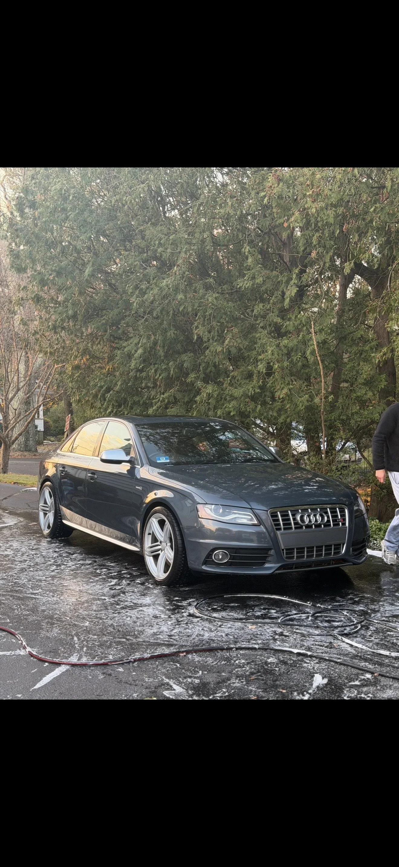 A black Audi sedan parked on a driveway being washed with foam, surrounded by trees and a person partially visible on the right side.