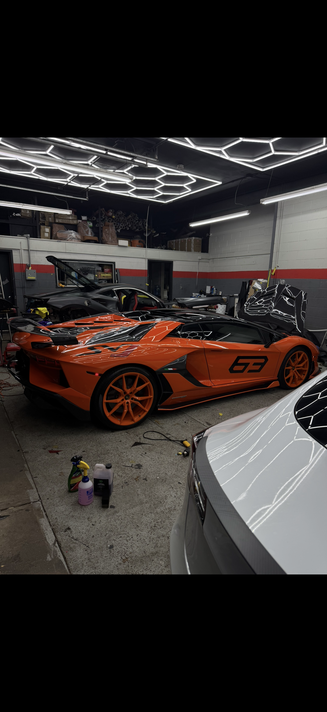 Orange sports car with racing number 63 in a garage with other vehicles, tools, and equipment.