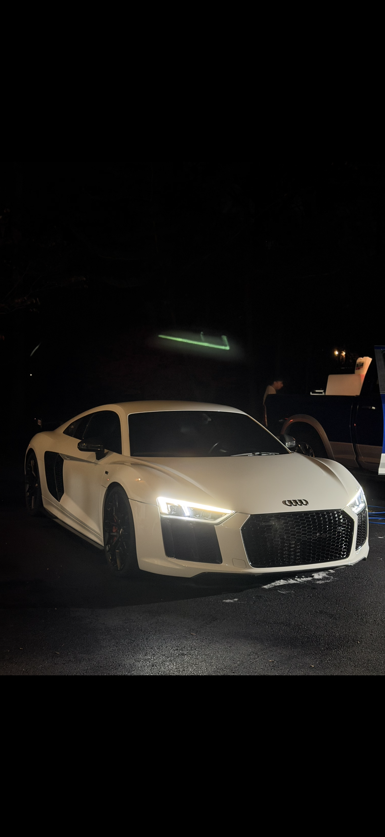 A white Audi R8 sports car parked at night with its headlights on, next to other vehicles in a parking lot.