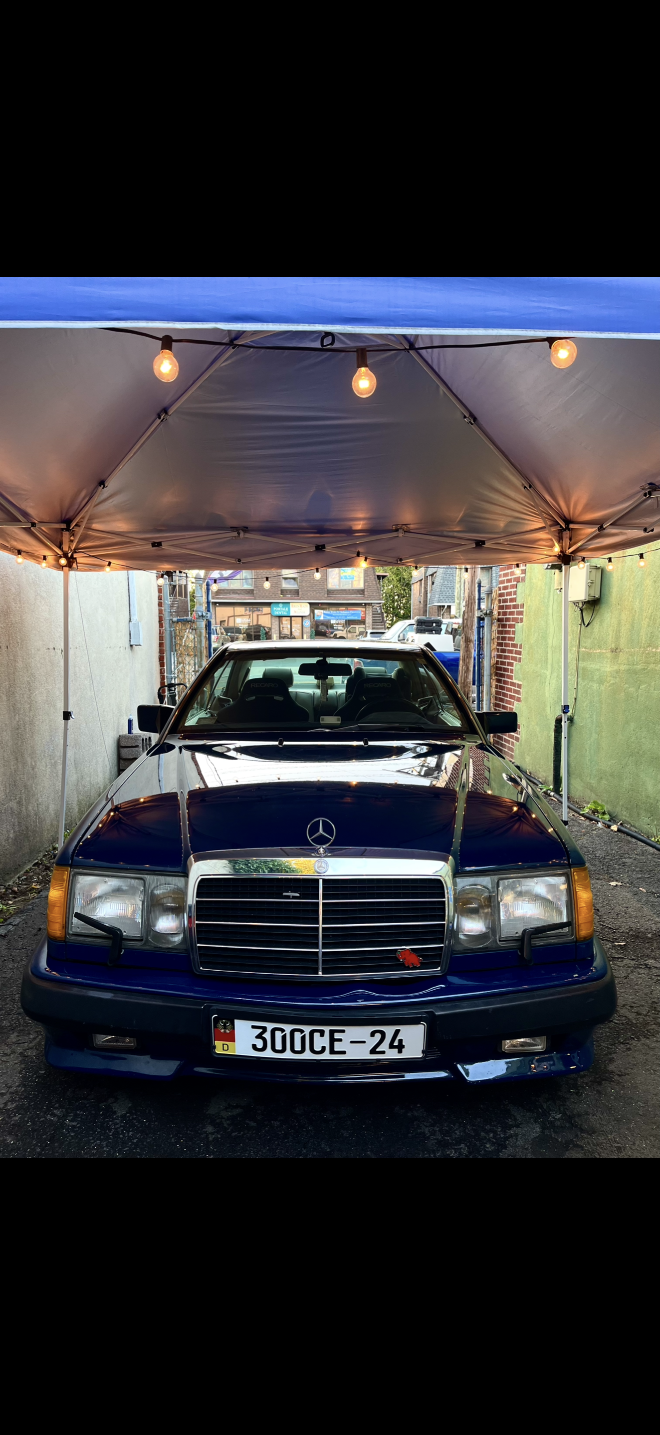 A blue vintage Mercedes-Benz car parked under a canopy with string lights, in an alleyway between buildings.