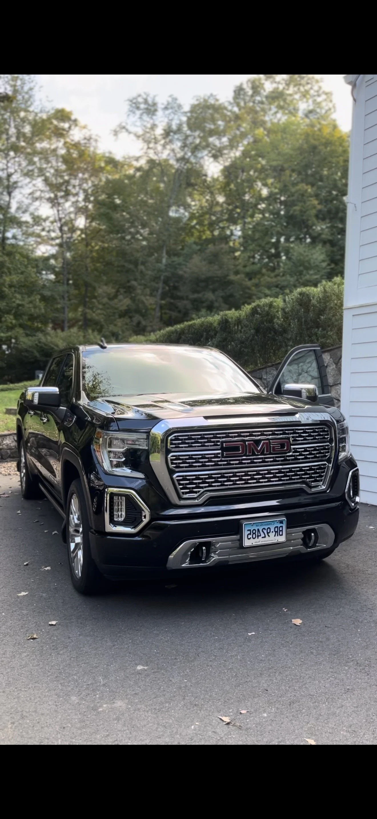 A shiny black GMC pickup truck parked outside near a white house, with one door open, surrounded by trees and bushes in the background.