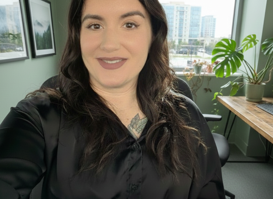 A woman with long dark hair, wearing a black satin blouse, smiling at the camera in an office with large windows, potted plants, and city buildings outside.