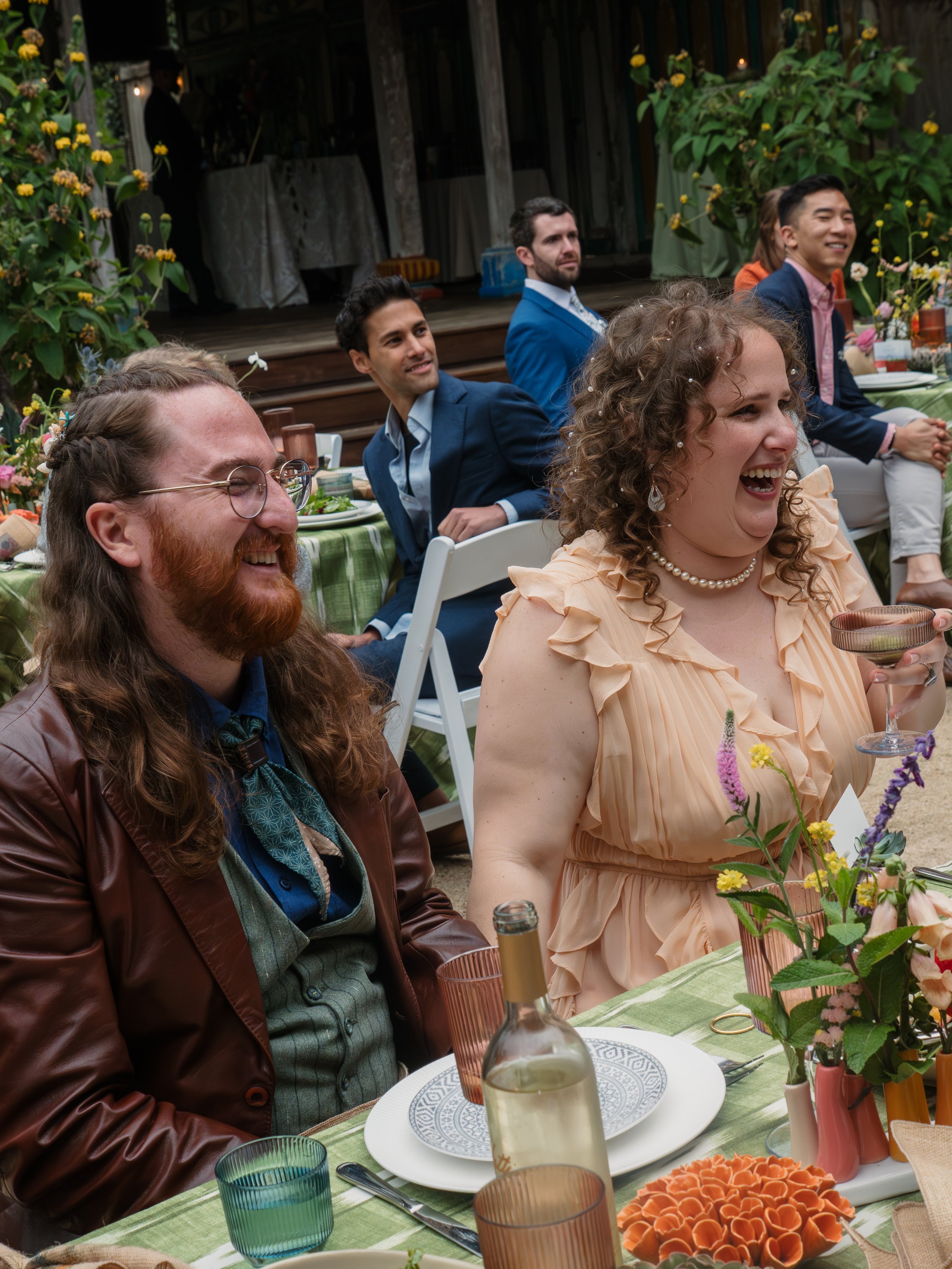 People sitting at a table decorated with flowers, enjoying a celebration outdoors, smiling and laughing.