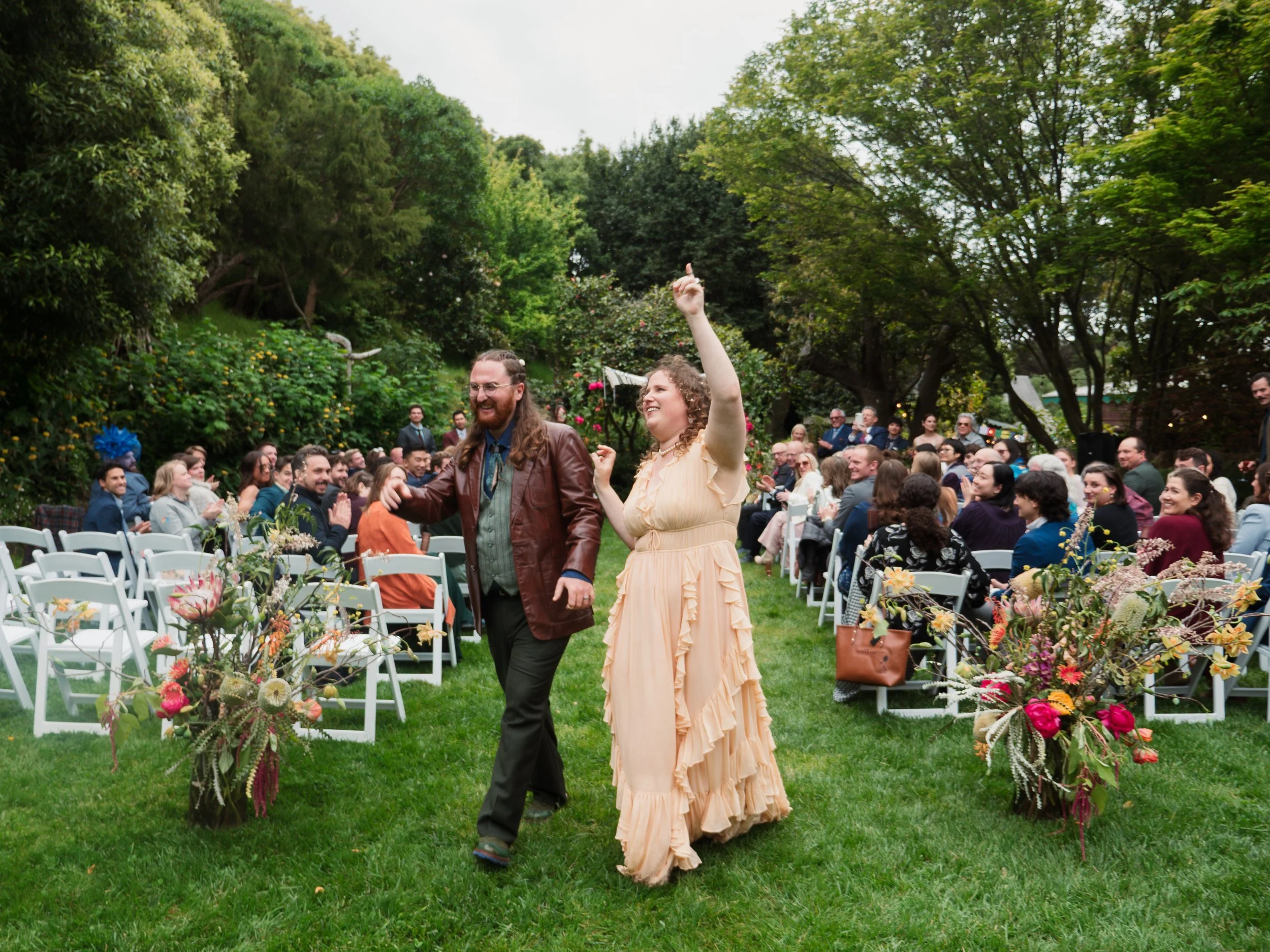 Two people, a man with long hair and a woman with curly hair, are walking together at their wedding, smiling and raising their hands. They are surrounded by seated guests in an elegant garden setting with trees and floral decorations.