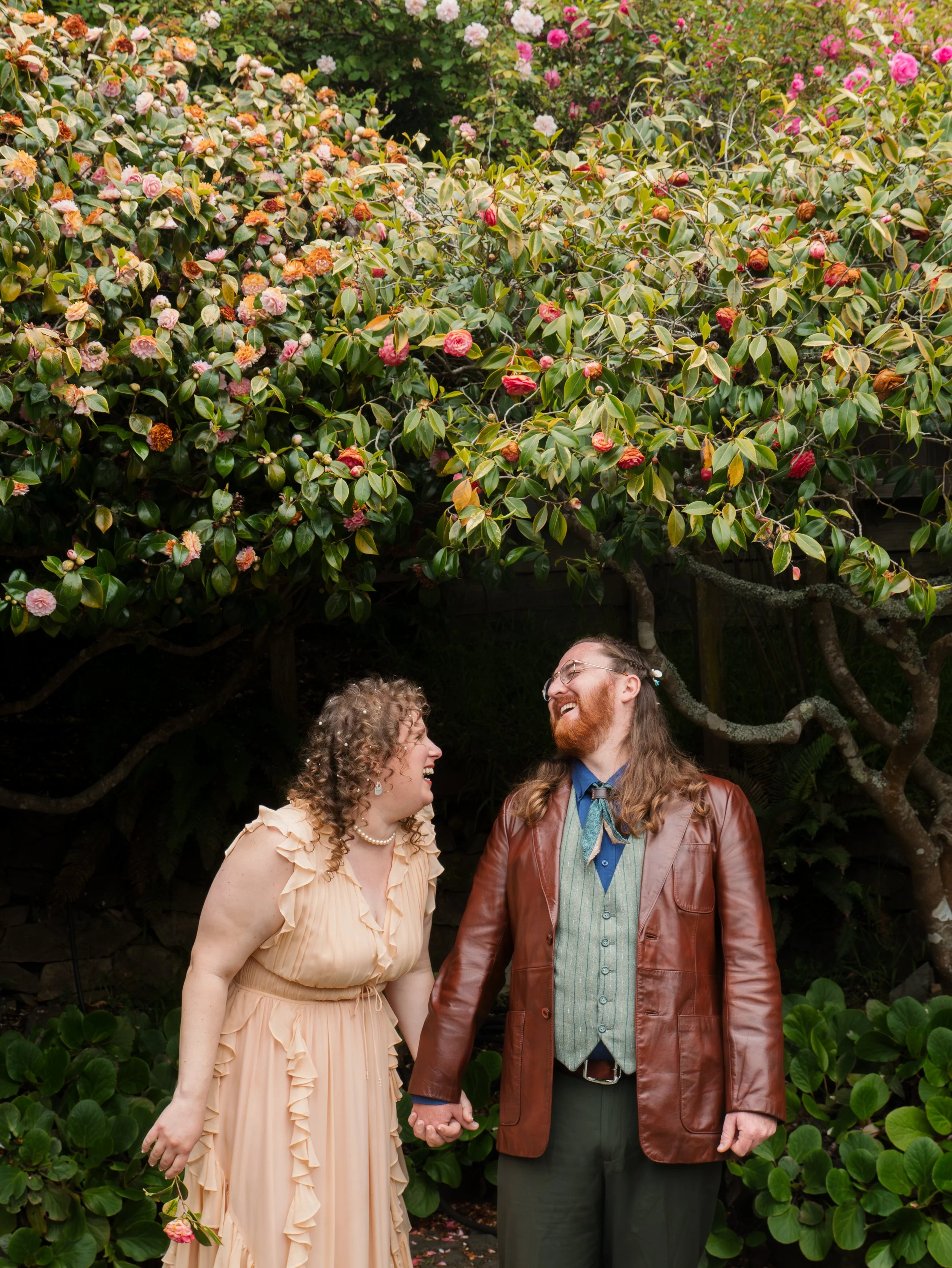 A happy couple holding hands and looking at each other in front of a lush bush with pink and white flowers.