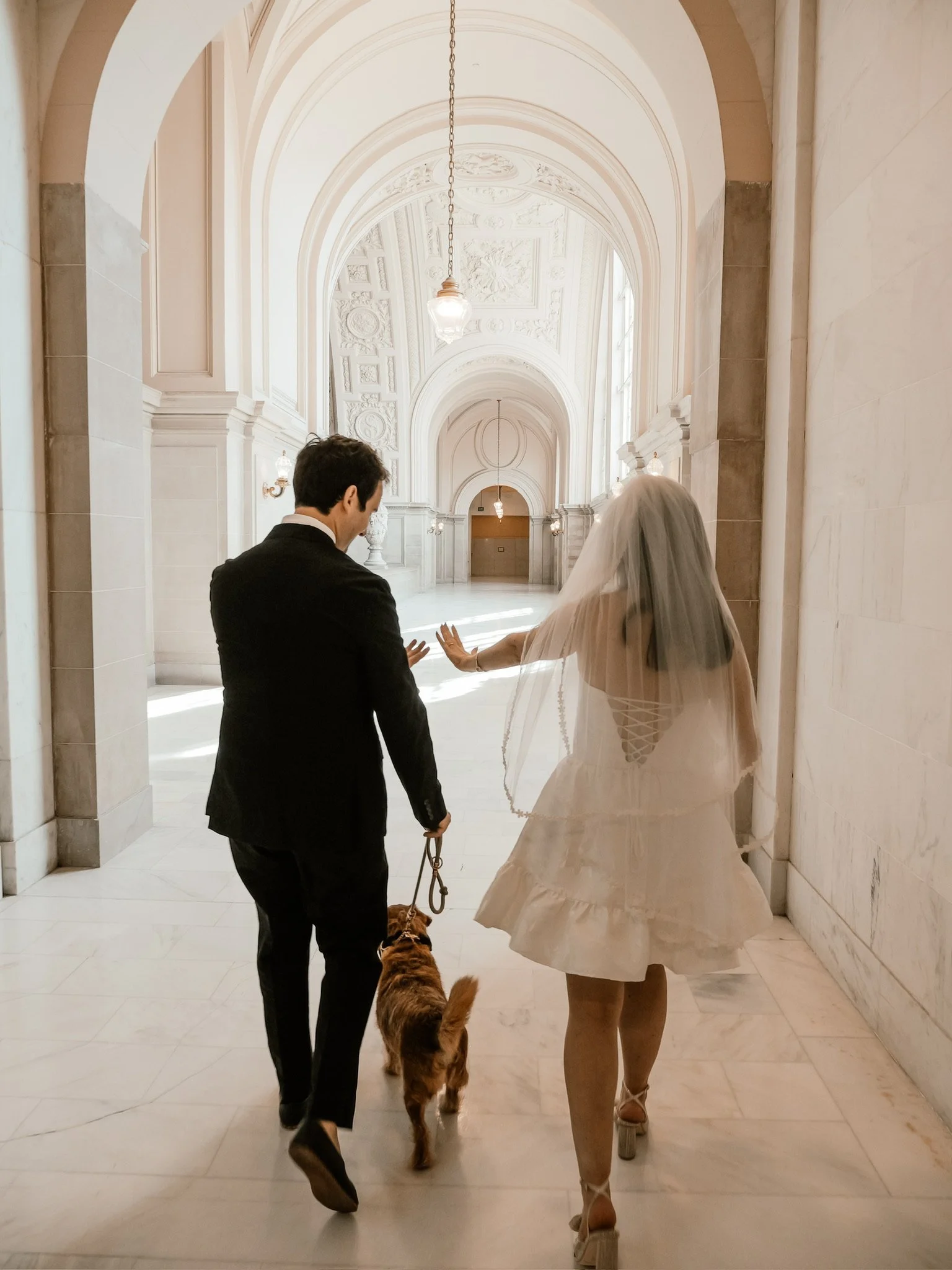 A bride and groom walking hand in hand through a grand hallway, accompanied by a woman and a dog, in an elegant wedding setting.