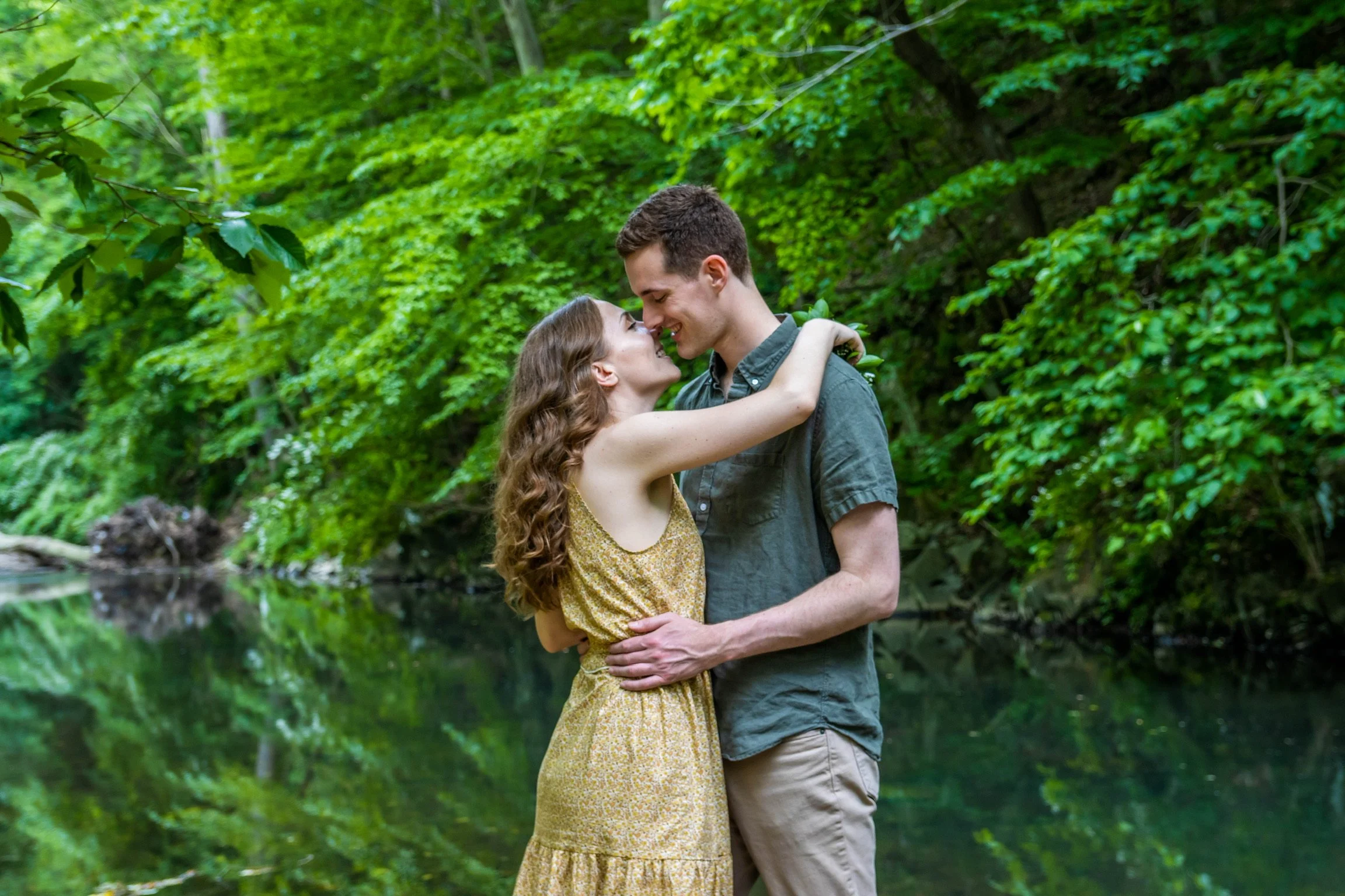 A young couple is embracing each other and smiling, standing outdoors near a river with lush green trees in the background.